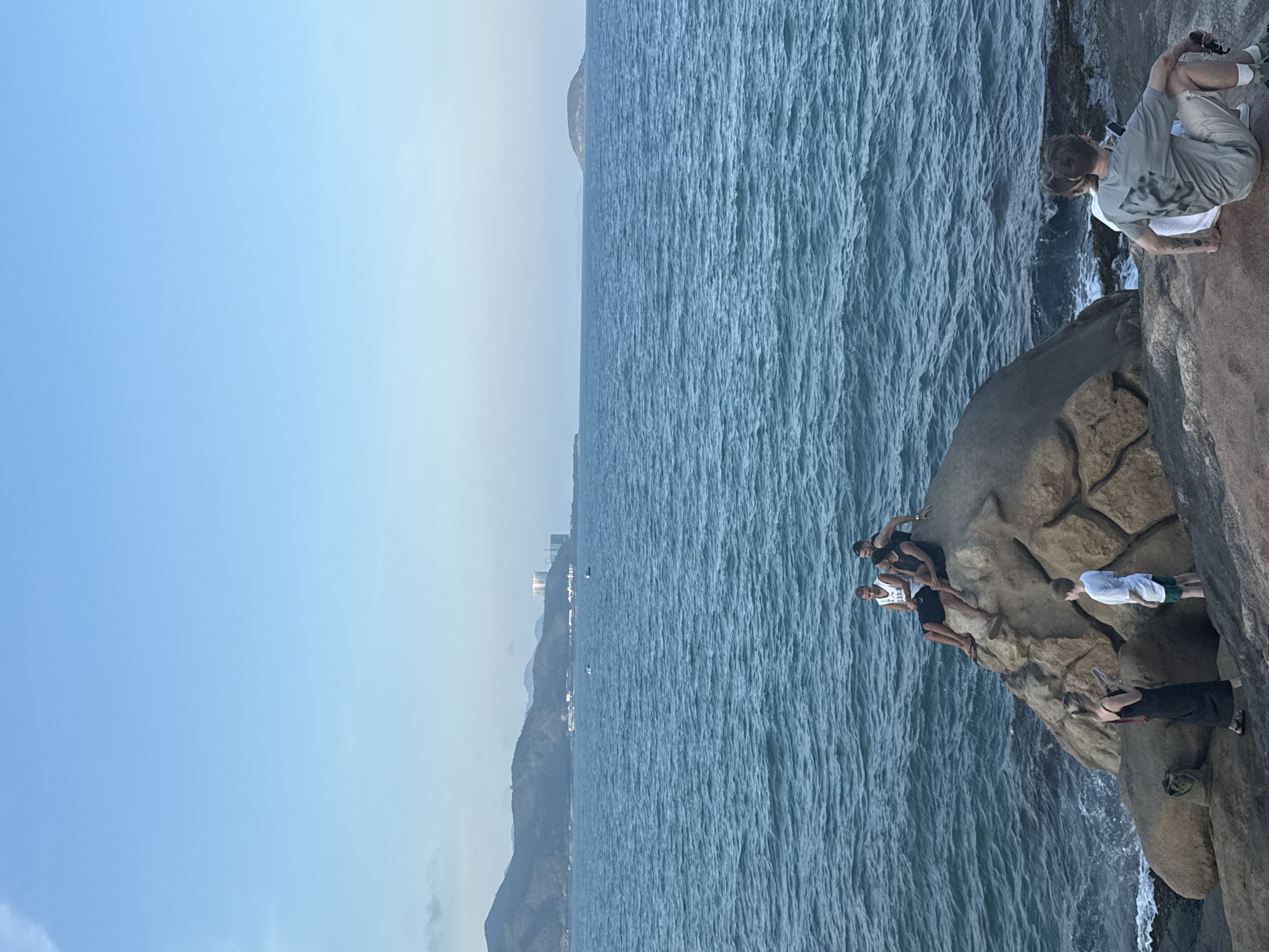 The image depicts a coastal scene at Bac Nha Trang Ward, Vietnam, with a group of people on a rocky outcrop overlooking the ocean. Six people are visible on the rocks; one man is sitting cross-legged on the rock with a camera in his hand, seemingly taking a photograph of the ocean. At least four other individuals are on top of the rock, with one person standing. The ocean extends towards the horizon. The sky is predominantly clear blue.