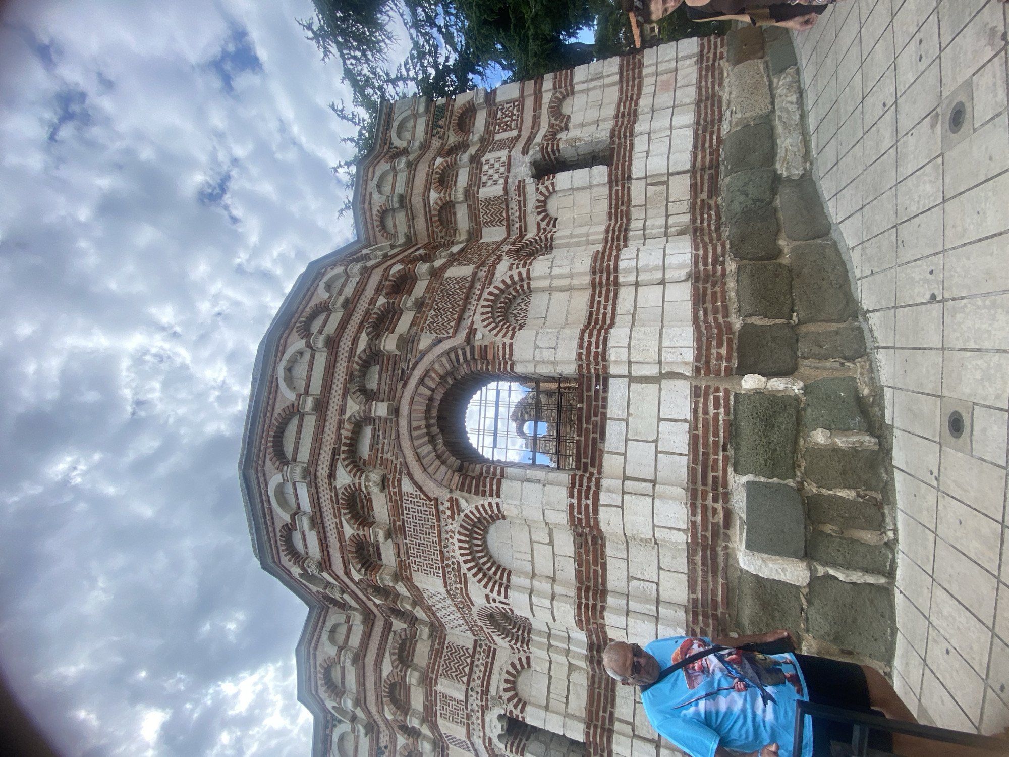 In Nesebar, Bulgaria, a man wearing a blue t-shirt with a graphic print and black shorts walks past a historic brick and stone building. The building exhibits intricate architectural details with alternating bands of red brick and lighter stone, adorned with arches and decorative patterns. A window with a metal grate reflects the cloudy sky and part of the building. A woman in a brown top is visible on the right side of the frame. The ground is paved with light-colored tiles. The sky is overcast with dramatic clouds.