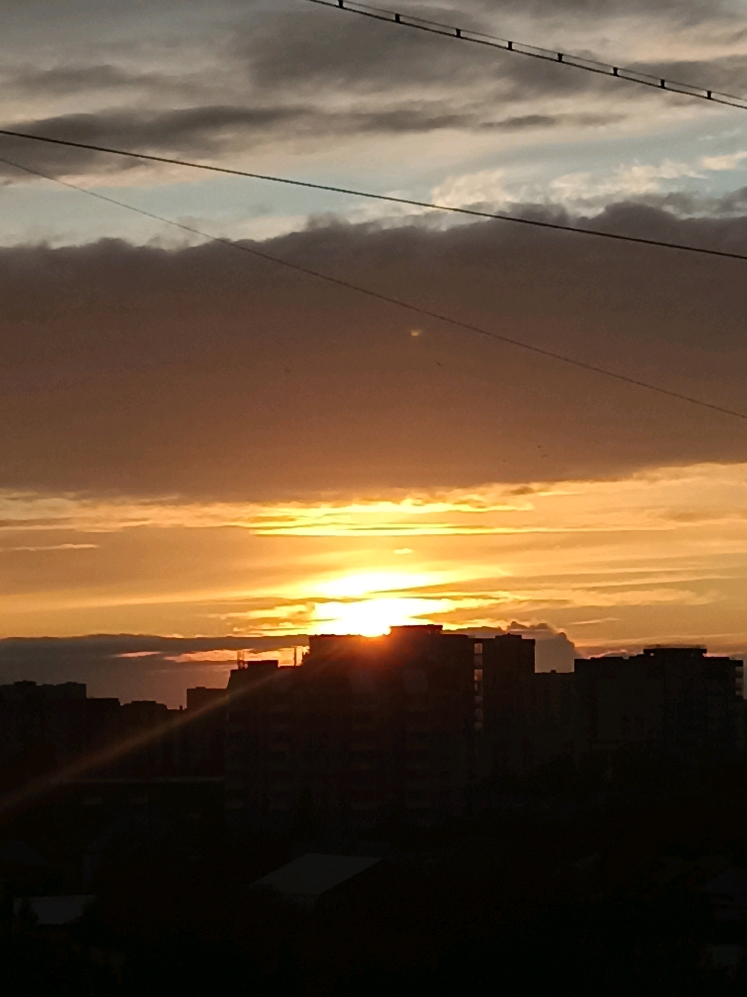 The image shows a sunset over a city skyline. The sun is low on the horizon, casting a warm orange glow across the sky. Clouds are scattered across the sky, with some tinged with pink and purple. The city skyline is silhouetted against the bright sky, with the shapes of buildings clearly visible. There are power lines running across the top of the image. The scene appears to be in a residential area, with apartment buildings dominating the skyline. This image was captured on June 11, 2024, at 9:01 PM in Novosibirsk, Russia.