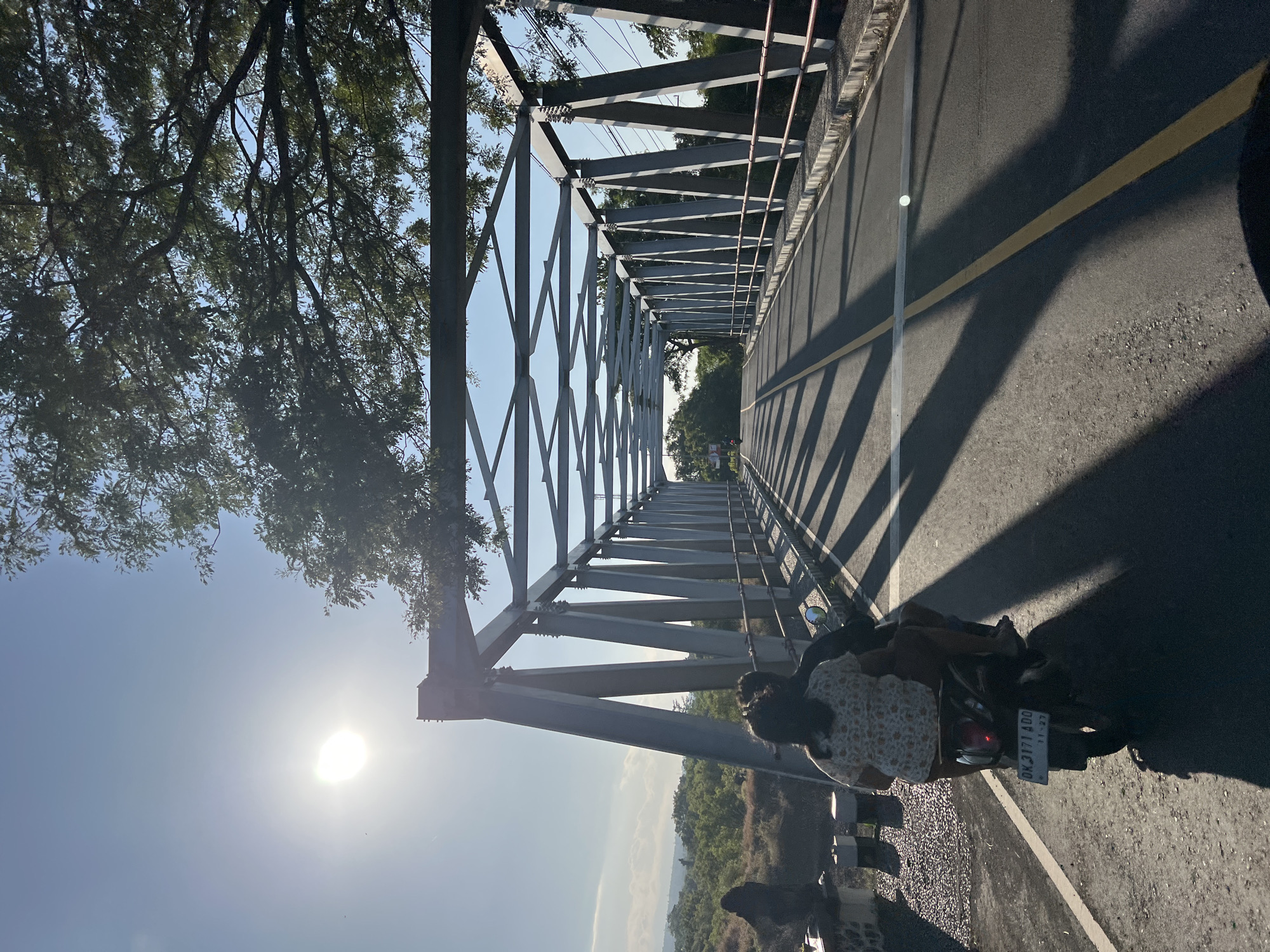 In Kubu, Indonesia, two individuals are riding a scooter across a metal truss bridge on a sunny day. The sun is bright, casting long shadows from the bridge's structure onto the asphalt road. The scooter has a license plate that reads "DK 3171 ADO". Trees line the sides of the road leading up to and over the bridge, with lush green foliage visible in the background. The bridge itself is a prominent feature, with its metallic beams creating a geometric pattern against the sky. The sky is clear and blue, with a few wispy clouds in the distance. The overall scene depicts a serene journey across a bridge in a tropical environment.