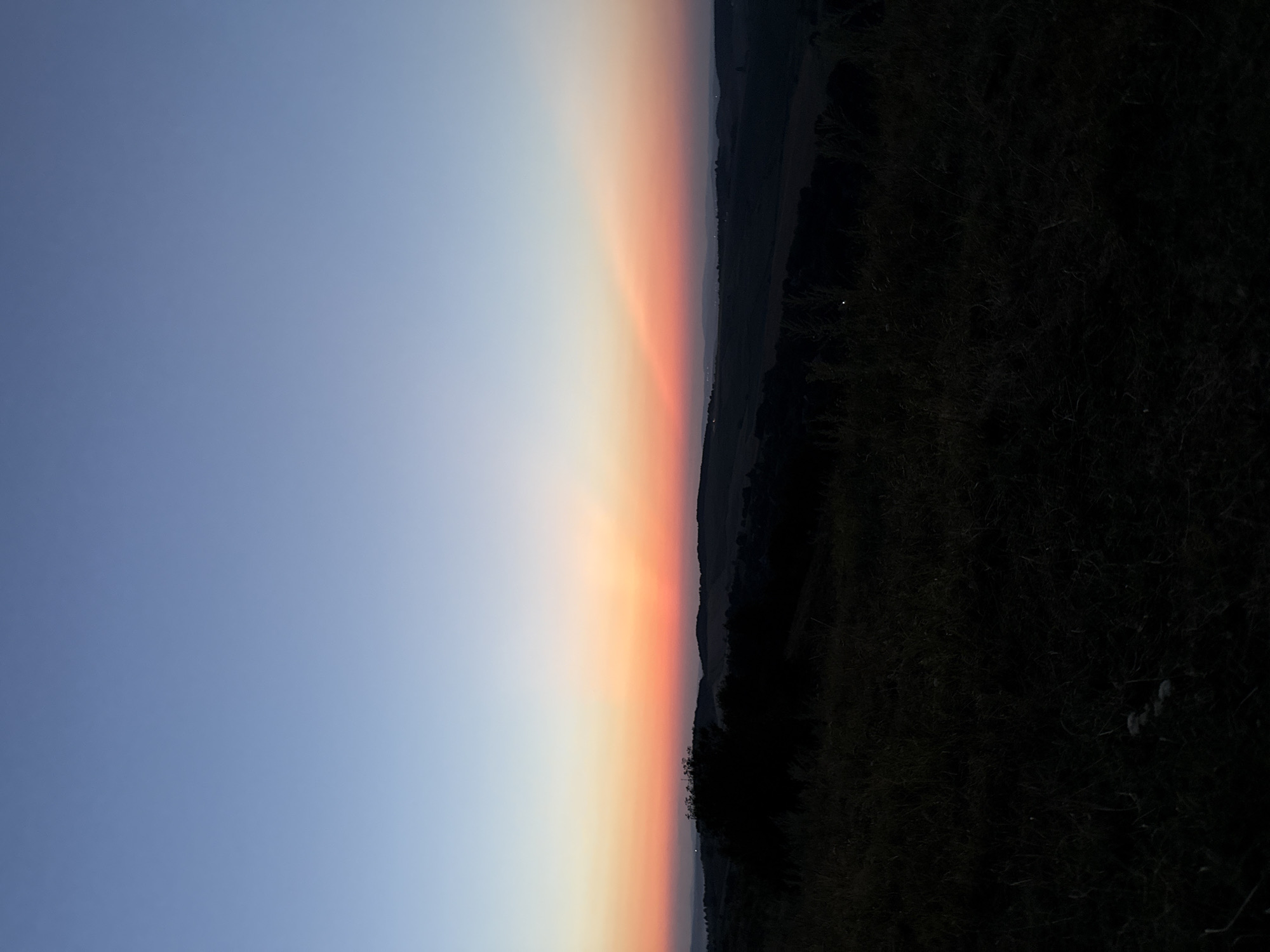 This is a landscape photograph taken at dusk or dawn. The sky transitions from a pale blue at the top to a gradient of orange and pink near the horizon. The horizon is marked by rolling hills covered in dark vegetation. The foreground consists of a dimly lit grassy area, with some faint white specks scattered throughout, possibly pebbles or dew. The overall scene is peaceful and serene, with no people or identifiable structures. The location is described as Unknown, Unknown.