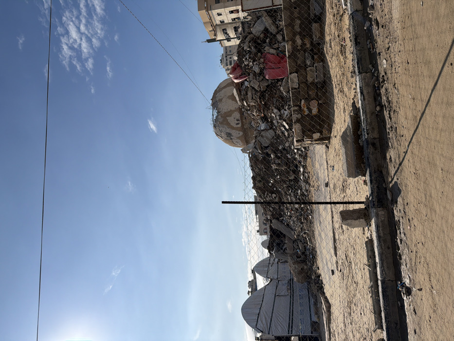 This image depicts a large demolition site in Gaza City, Palestinian Territory, under a bright, mostly clear blue sky. In the foreground, sandy ground with sparse debris is present, bordered by a broken concrete curb and a chain-link fence, which includes a vertical black metal post. The midground is dominated by a substantial pile of building rubble, composed of fragmented concrete, rebar, and other construction debris. Perched on top of this rubble pile, slightly to the right of center, is a partially destroyed, light-colored dome structure exhibiting visible damage and exposed internal materials. Two large, prominent red tarpaulins or sacks are visible on the debris to the right of the dome. To the far left, behind the fence and partially obscured by the rubble, are several white, arched, tent-like structures with dark horizontal bands. The background reveals an urban skyline featuring multiple light-colored multi-story buildings. Utility lines stretch horizontally across the upper portion of the frame, and a bright sun flare is visible in the upper left corner. No people are visible in the scene.