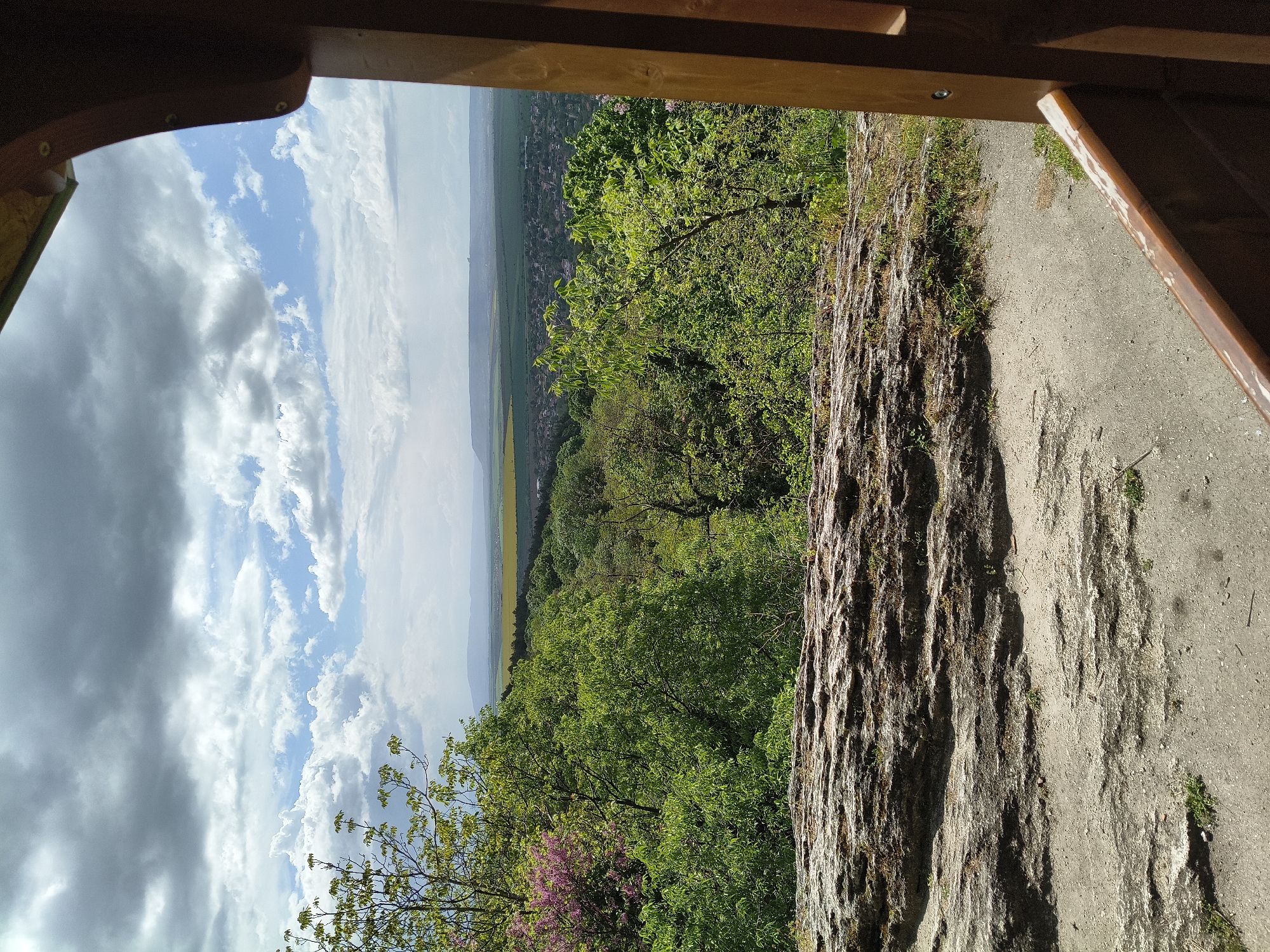 A view from an elevated wooden structure overlooking a landscape on a partly cloudy day. A rocky outcropping is visible in the foreground, leading to a forest of green trees. Beyond the trees, a distant cityscape or town is visible, with agricultural fields in between. The sky has a mix of dark and light clouds. The image is shot from a first-person perspective, looking out from within the wooden structure.
