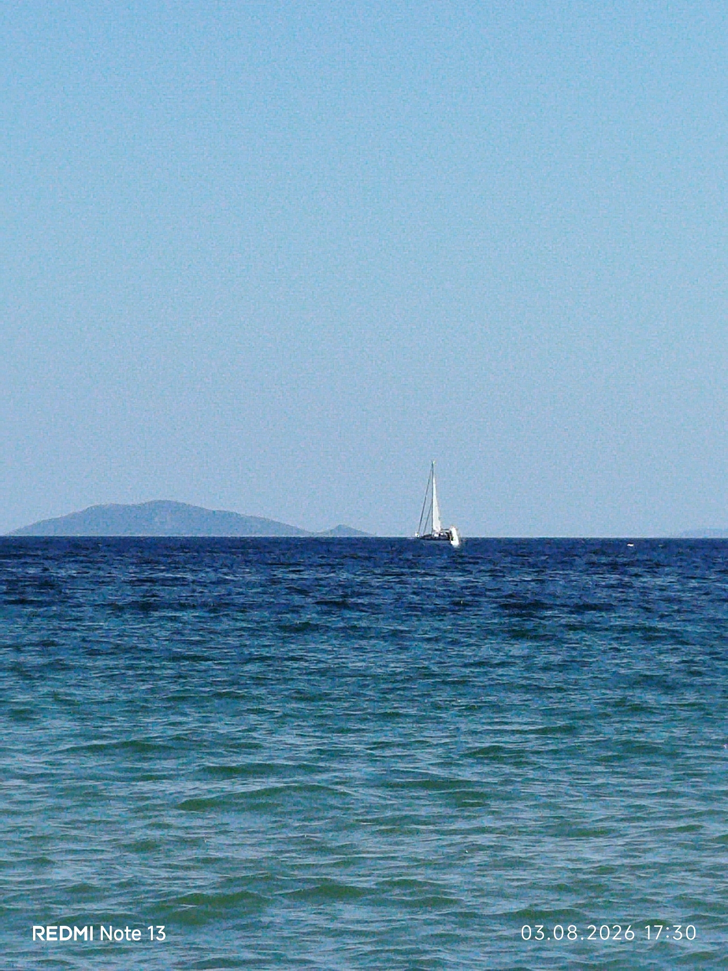 The image shows a wide expanse of the Aegean Sea under a clear blue sky. In the distance, a faint outline of a mountainous island can be seen. A white sailboat with its sails raised is positioned in the middle of the sea, appearing small against the vastness of the water. The water itself is a deep blue, transitioning to a lighter turquoise closer to the foreground, with small waves creating a textured surface. The bottom left of the image displays the text "REDMI Note 13", and the bottom right shows "03.08.2026 17:30", indicating the date and time the photo was taken. This scene is representative of the coastal areas within Sithonia Municipal Unit, Greece, known for its beautiful beaches and clear waters.