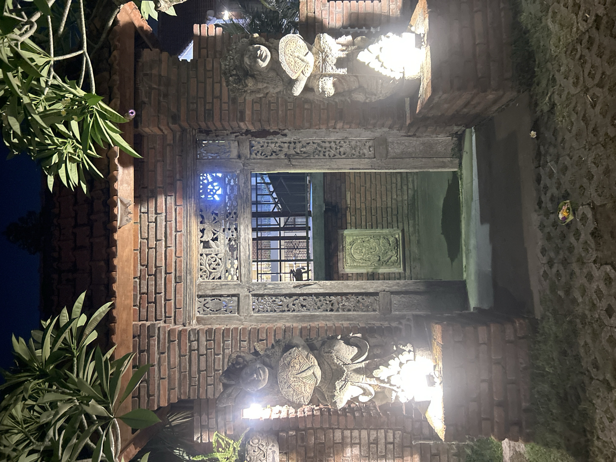 This is a photograph taken at night of an entryway in North Kuta, Indonesia. The entryway is framed by brick pillars and features ornate wooden carvings. On either side of the entrance, there are illuminated stone statues of figures holding fans. Behind the statues, a brick building is visible, with a metal fence and stairs leading upwards. Lush green foliage hangs from above, casting shadows. The ground is paved with interlocking stones and patches of grass. The scene is lit by artificial lights, giving it a warm glow.