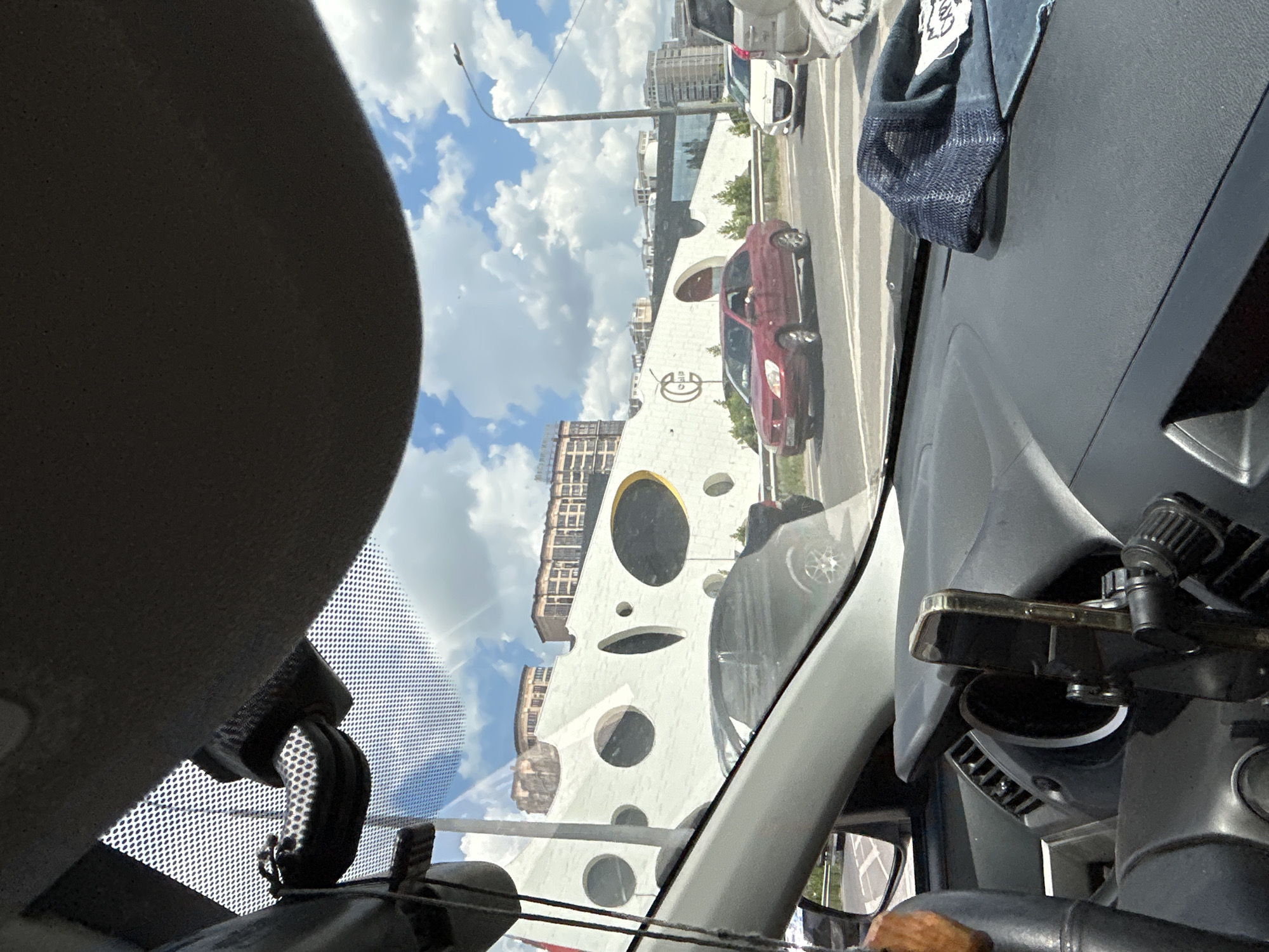 The image is taken from the interior of a car, looking out through the windshield at a street scene. The car's dashboard is visible in the foreground, with a smartphone mounted on a holder. A dark blue mesh cap rests on the dashboard. Outside, traffic is moving on a road with several cars visible. A distinctive white building with large, circular cutouts and dark interiors dominates the background. This building is situated in the city of Orlovskii, Russia. The sky is blue with scattered white clouds.