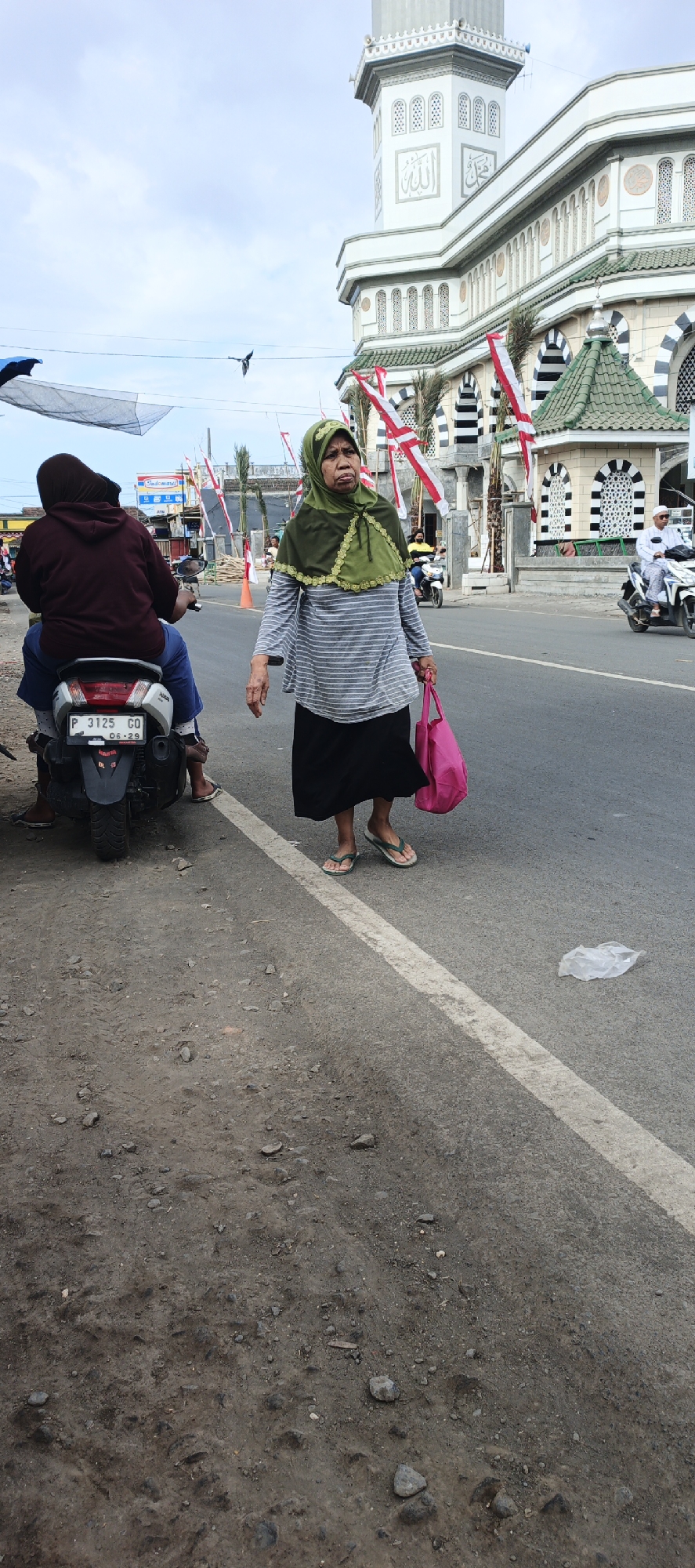 The image captures a street scene in Puger, Indonesia, with a mosque prominently featured in the background. A woman wearing a green hijab and a striped long-sleeved shirt, along with a black skirt, is walking on the right side of the road, carrying a pink bag. She is wearing flip-flops. On the left side of the road, a person sits on a motorcycle, facing away from the camera, wearing a maroon hooded jacket and blue shorts. Their feet, clad in flip-flops, are visible. The motorcycle has a license plate that reads "P 3125 GO". The road is paved, and the surrounding environment appears to be a town or city area, with flags and palm trees visible in the background near the mosque. The sky is cloudy.