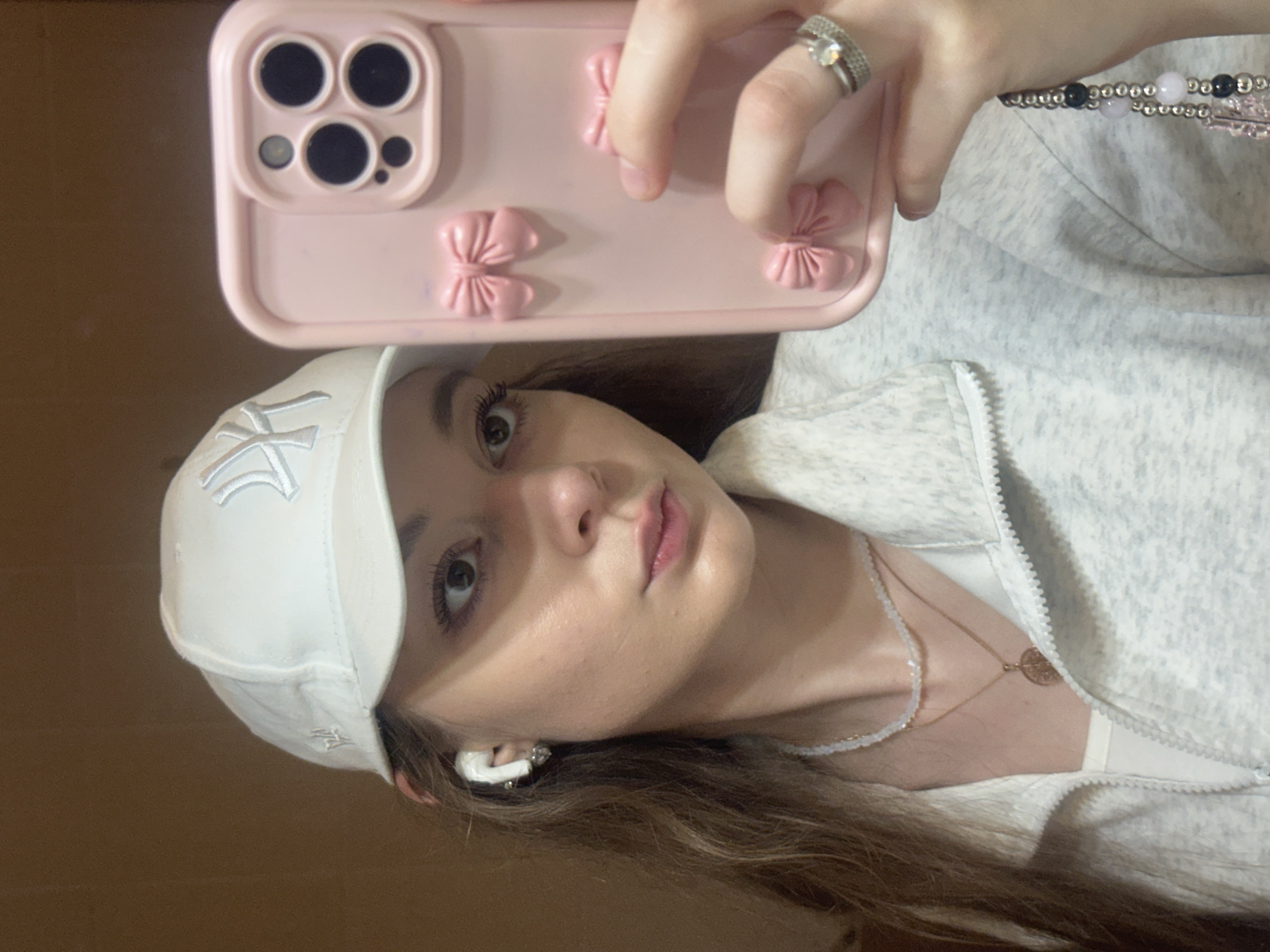 A young woman is taking a selfie in what appears to be a home environment. She is wearing a white baseball cap with a New York Yankees logo, wireless earbuds, a light grey zip-up jacket over a white top, and two necklaces. She is also wearing rings on her right hand, which is holding up a pink phone case adorned with bows. Her long, brown hair is visible. The background is a plain, brown wall. The provided crops focus on her face, accessories, and the phone case. There is no indication of the location being in Kazan, Russia.