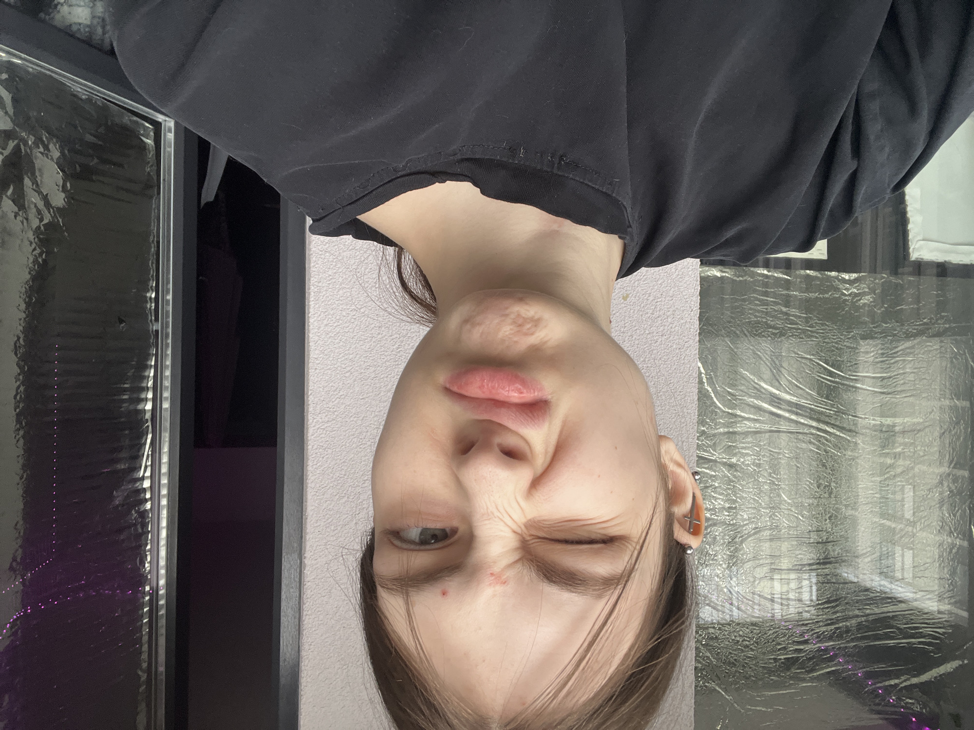 An upside-down portrait features a young person with light skin and dark hair, wearing a black t-shirt. The person is making a playful or humorous facial expression, with their right eye (viewer's left) wide open and looking towards the camera, while their left eye (viewer's right) is tightly winked or squinted, creating noticeable wrinkles around it. Their lips are pursed, adding to the distorted, comical look. Small red blemishes are visible on the forehead and chin. The person has multiple piercings on their right ear, including one shaped like a cross and several stud piercings along the helix.
The background consists of a light-colored, textured wall behind the person's head. To the person's left and right sides (which would be the top and bottom of the upright image), there are large areas covered with crinkled, reflective material, possibly plastic film or foil, reflecting light and displaying hints of purple luminescence in some areas. Dark, rectangular elements, possibly doorways or furniture, are partially visible in the deeper background. The setting appears to be an indoor environment, with no discernible time of day or weather conditions. No text is visible in the image. The provided context indicates the location is Saint Petersburg, Russia.