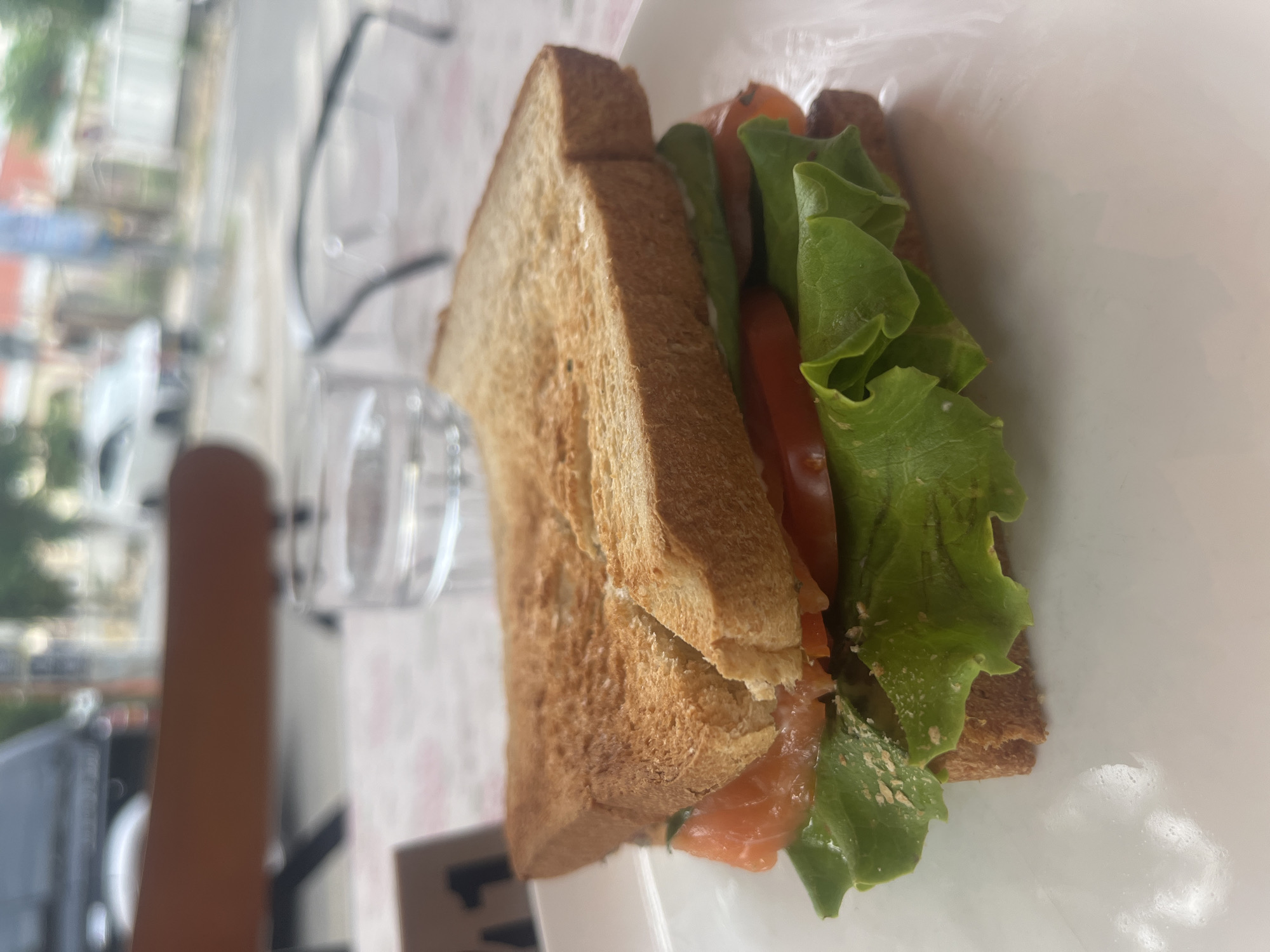 An outdoor cafe in Da Nang, Vietnam, features a salmon sandwich on a white plate. The sandwich has toasted bread, lettuce, tomato, and salmon. A glass of water and a pair of glasses are on the table behind the sandwich. In the background, cars and buildings are visible.