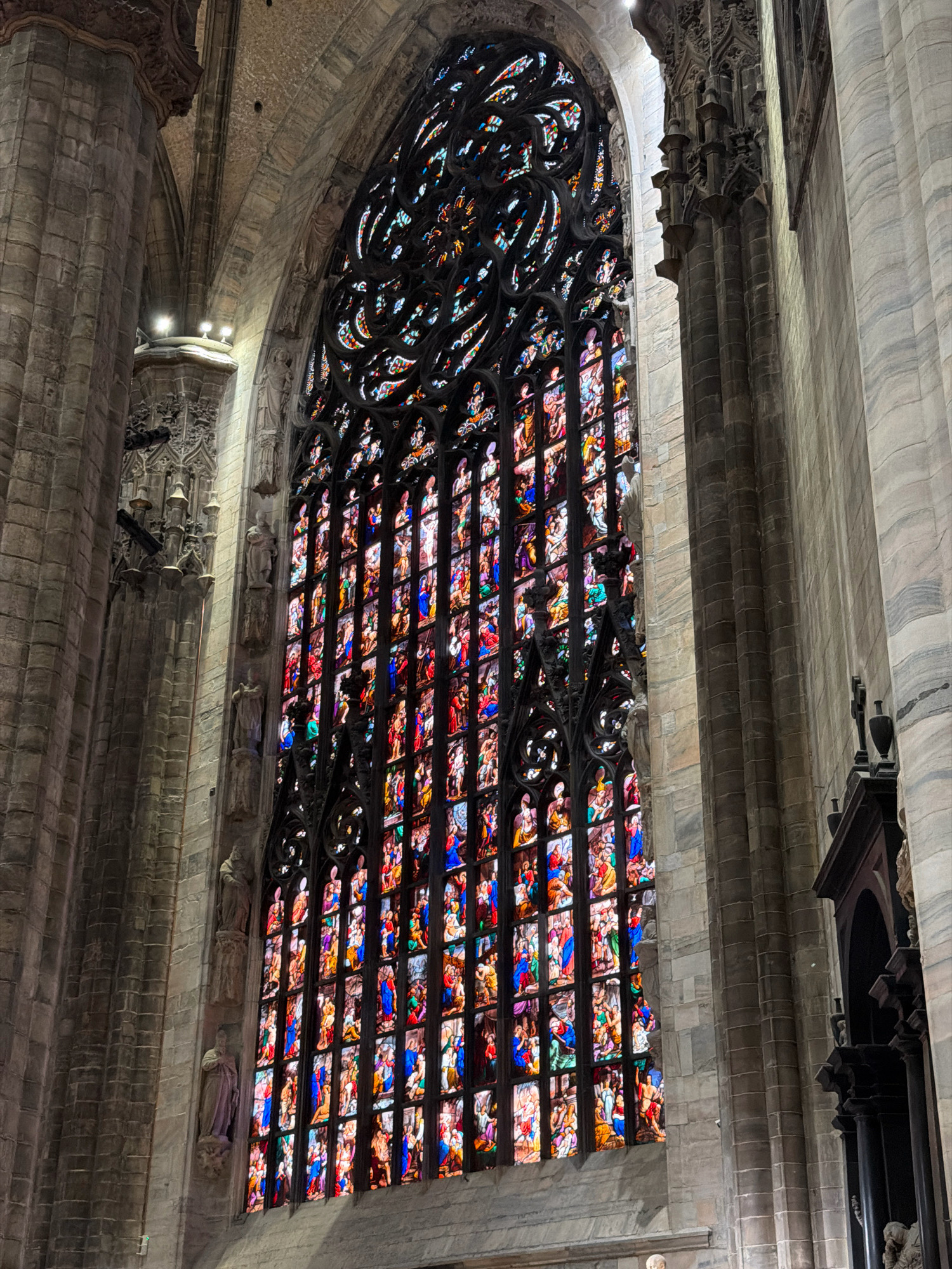 The image shows a large, ornate stained-glass window within a grand Gothic-style cathedral. The window is tall and arched, filled with intricate patterns and numerous individual panels, each depicting colorful scenes with human figures. The glass glows with reds, blues, purples, greens, and yellows, illuminated from behind by unseen light. The architectural surroundings are made of stone, with massive columns and vaulted ceilings visible. There are also carved statues of figures in niches along the walls. The overall impression is one of immense scale, intricate artistry, and religious grandeur, characteristic of a significant European cathedral, likely the Duomo di Milano (Milan Cathedral) given its famous stained-glass windows.