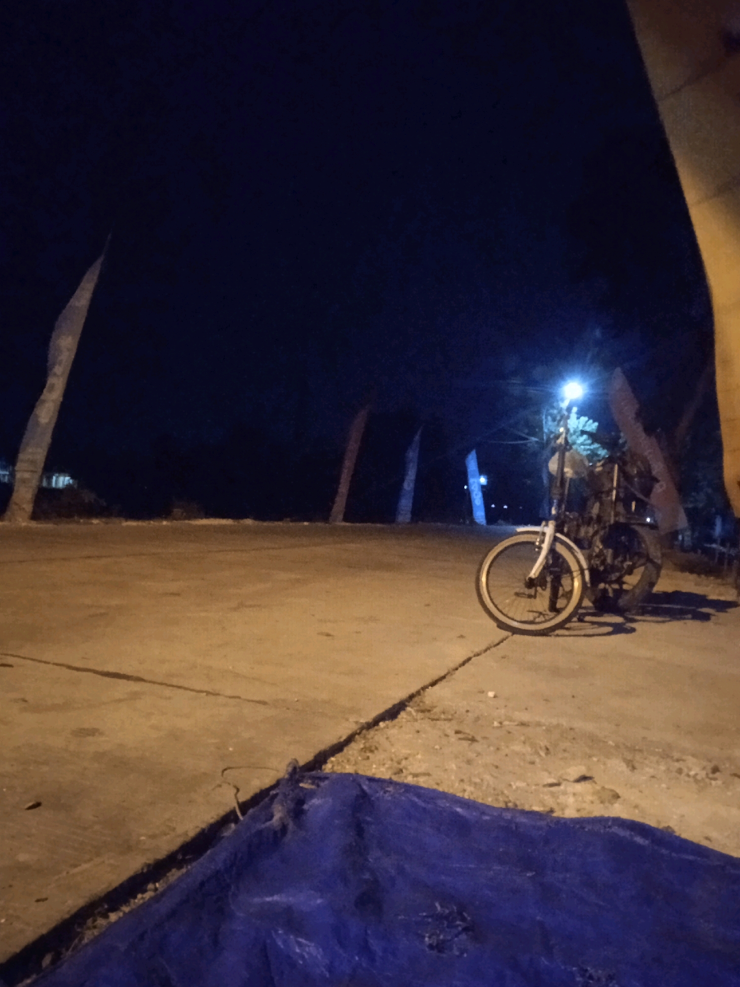 A nighttime scene in the city of Sumodikaran, Indonesia, shows a paved surface and a dirt area. In the foreground, a large blue tarp is spread out. Further back, a bicycle is parked under a streetlamp, which illuminates the vehicle and the immediate surroundings. The background features several tall, thin, tree-like structures, likely palm trees, and a dark sky, suggesting it is late at night. The overall impression is one of a quiet, sparsely lit outdoor area.