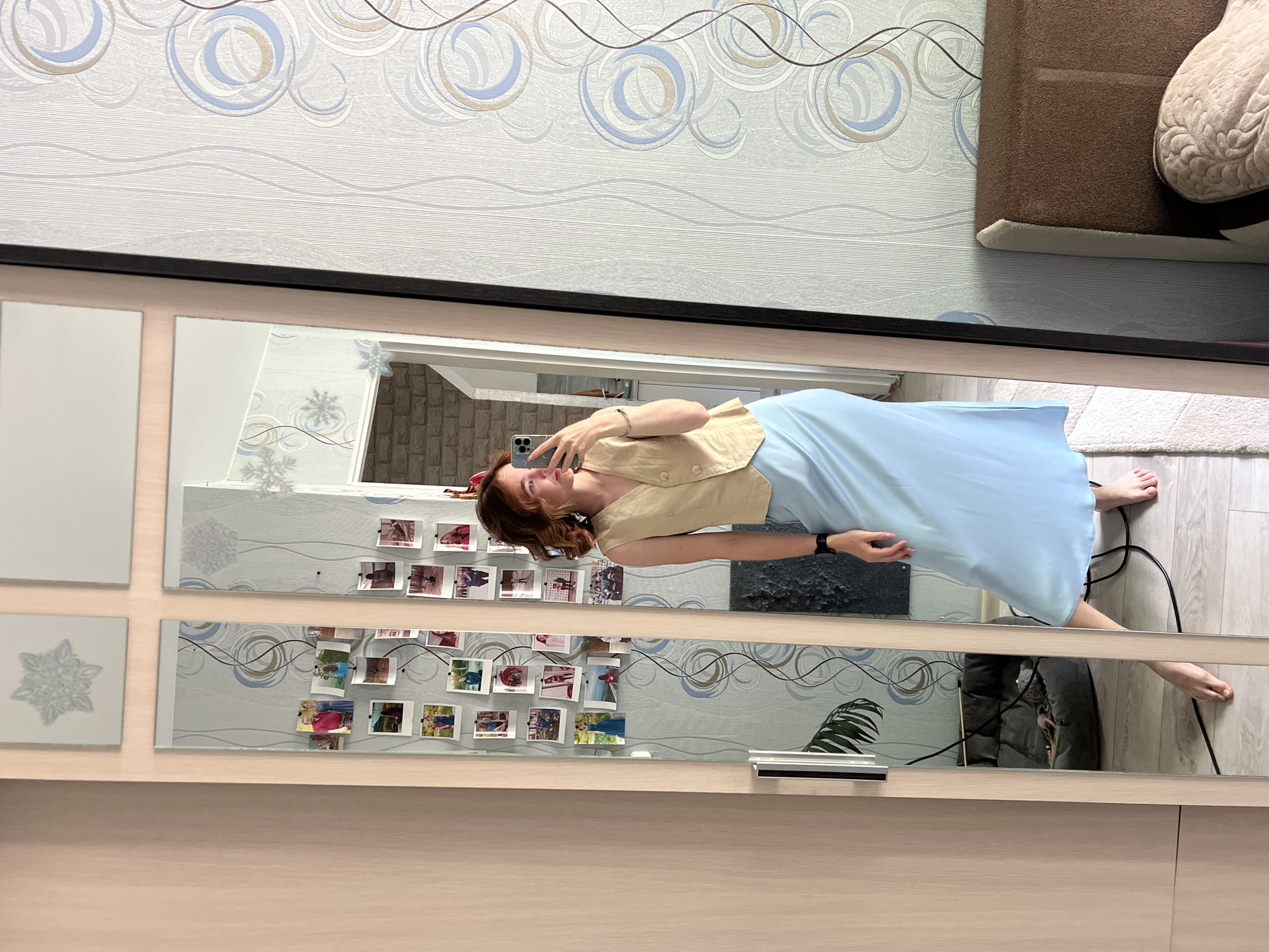A young woman with shoulder-length red hair stands in front of a full-length mirror, taking a selfie with her phone. She is wearing a light beige sleeveless vest over a pale blue slip dress. The mirror is part of a wardrobe, and it reflects the room behind her. The room's wallpaper features a swirling pattern of light blue, beige, and grey circles. Several small framed photos are pinned to the mirror frame. In the reflection, a dark gray pet bed with a small dog is visible, along with a plant in a pot. The woman's bare feet are visible in the foreground, standing on a white rug. The image appears to have been taken indoors, likely in a bedroom. The subject is posing in her apartment in the city of Chaikovsky, Russia.