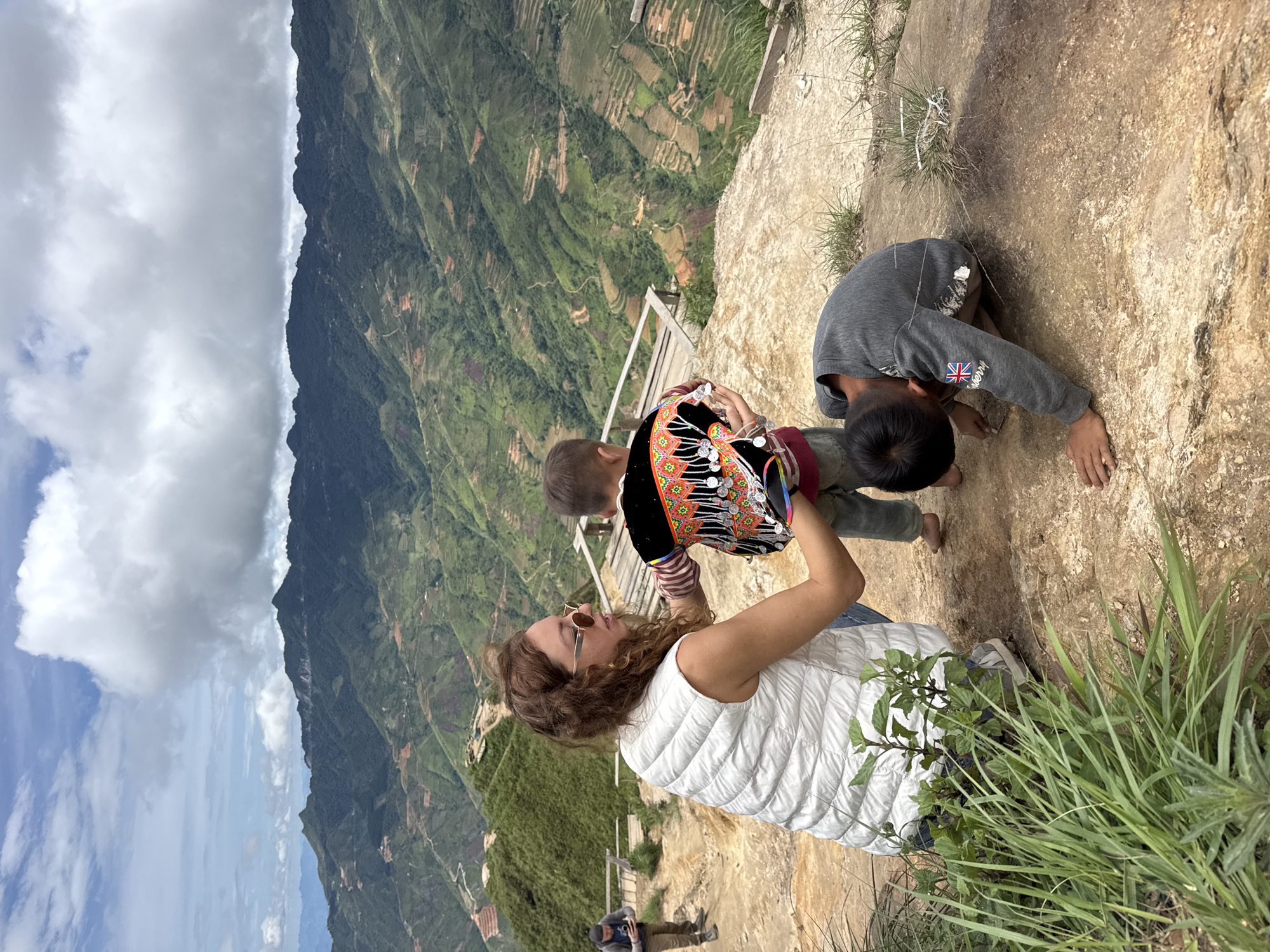 A woman with curly brown hair and sunglasses kneels on a rocky surface, wearing a white vest and blue jeans, and adjusting the traditional colorful jacket of a young child. The child, facing away from the camera, wears the jacket over a red shirt and denim shorts, with bare feet. Another child, in a gray sweatshirt with a Union Jack patch on the sleeve and the word "berry" below it, kneels on the ground, looking down. In the background, a man in a dark hat and jacket stands on a wooden walkway, looking at his phone. The landscape is mountainous with terraced hillsides in various shades of green, indicative of the rice paddies common in rural Vietnam. The sky is partly cloudy with large white clouds. The location is the picturesque mountainous region of Bắc Yên, Vietnam, known for its stunning natural beauty and ethnic minority villages.