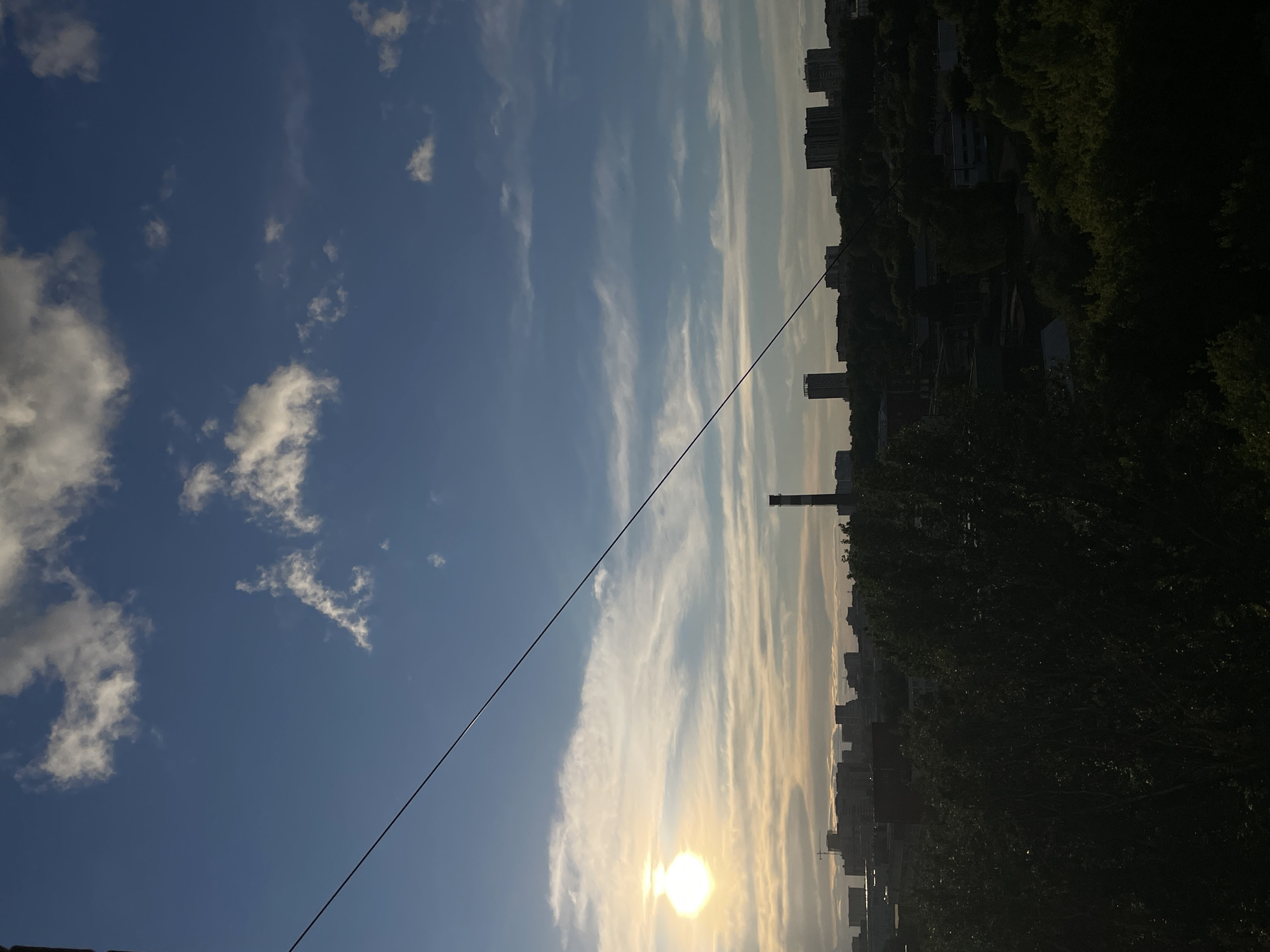 The image shows a view towards the sky and cityscape, likely in Izhevsk, Russia. A single power line is prominently angled from the bottom left to the upper right. The sky has patches of blue interspersed with light, streaky clouds. The sun is visible near the horizon, creating a bright, diffused glow. On the right side of the frame, there is a dense cluster of buildings and trees, suggesting an urban area bordering a park or wooded area. A tall, thin smokestack is also discernible amidst the buildings. The overall composition is unbalanced, with the sky and sun being the dominant elements, with a dark and somewhat obscured cityscape on the right.