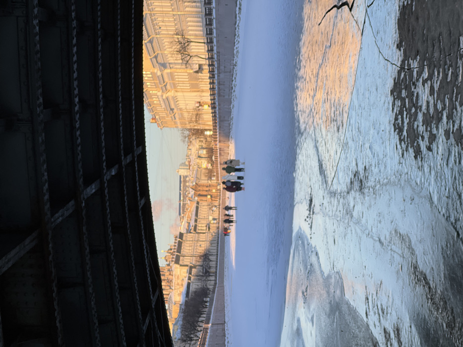 The image depicts a winter scene on the Griboyedov Canal in Saint Petersburg, Russia, viewed from directly underneath a bridge, looking eastward. The top portion of the frame is occupied by the dark, structural underside of the bridge, revealing its metal girders and rivets.
In the immediate foreground, the frozen surface of the canal displays a mix of textured ice, slush, and dark water, reflecting the soft light. Further into the midground, the canal is uniformly covered in white snow and solid ice. A group of approximately ten to fifteen individuals, dressed in various winter coats, hats, and scarves, are walking across the frozen canal surface. They are dispersed, forming two main clusters, and appear to be moving generally towards the distant right side of the frame.
The banks of the canal are lined with snow-covered ground and bare trees. On the left bank, a series of classical European buildings with intricate facades and multiple stories are visible, bathed in the warm, low light of the sun. Further in the distance on this side, the golden domes and architectural details of what is identifiable as the Church of the Savior on Spilled Blood are prominent. A faint wisp of white smoke or steam rises into the cold air from a distant building. On the right bank, a large, stately building with numerous windows and classical detailing stands brightly illuminated by the sun. The sky above is a pale, clear blue, transitioning to a warm, golden hue near the horizon where the sun is low. The scene indicates a cold, clear winter day.