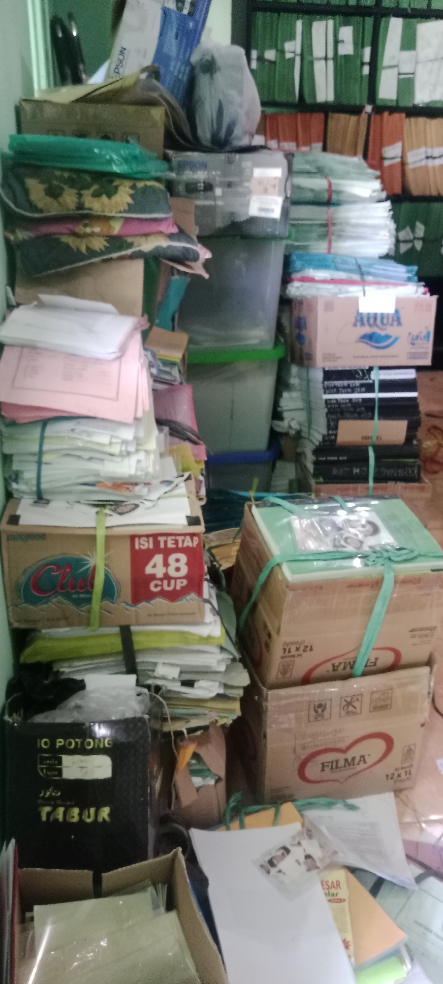 The image depicts a cluttered storage area, possibly a back office or stockroom, within a business located in Tuban, Indonesia. The environment is characterized by stacks of boxes, files, and miscellaneous items piled high, suggesting a lack of organization or a recent influx of inventory.
In the foreground, a black box with yellow lettering reads "IO POTONG TABUR" and appears to be a product container. To its right, several cardboard boxes are stacked, one prominently displaying the brand "FILMA" with "12 x 1L (pouch)" printed on it. Another box, lower down, shows an image of a red heart and the brand "FILMA" again. Above these, a box labeled "Club" with "ISI TETAP 48 CUP" indicates a product containing 48 cups. Further to the left, a stack of papers and folders is visible, with some bound by green string.
In the background, numerous folders are neatly organized on shelves, predominantly in green, orange, and yellow hues. There are also several large plastic storage bins, one appearing to hold more items. A printer box labeled "EPSON" is also visible among the clutter. The overall impression is one of a busy, functional space where storage is at a premium. No individuals are present in the image, and no actions or interactions are depicted. The lighting is somewhat dim, with a slightly greenish tint, possibly from overhead fluorescent lights. The floor appears to be tiled.