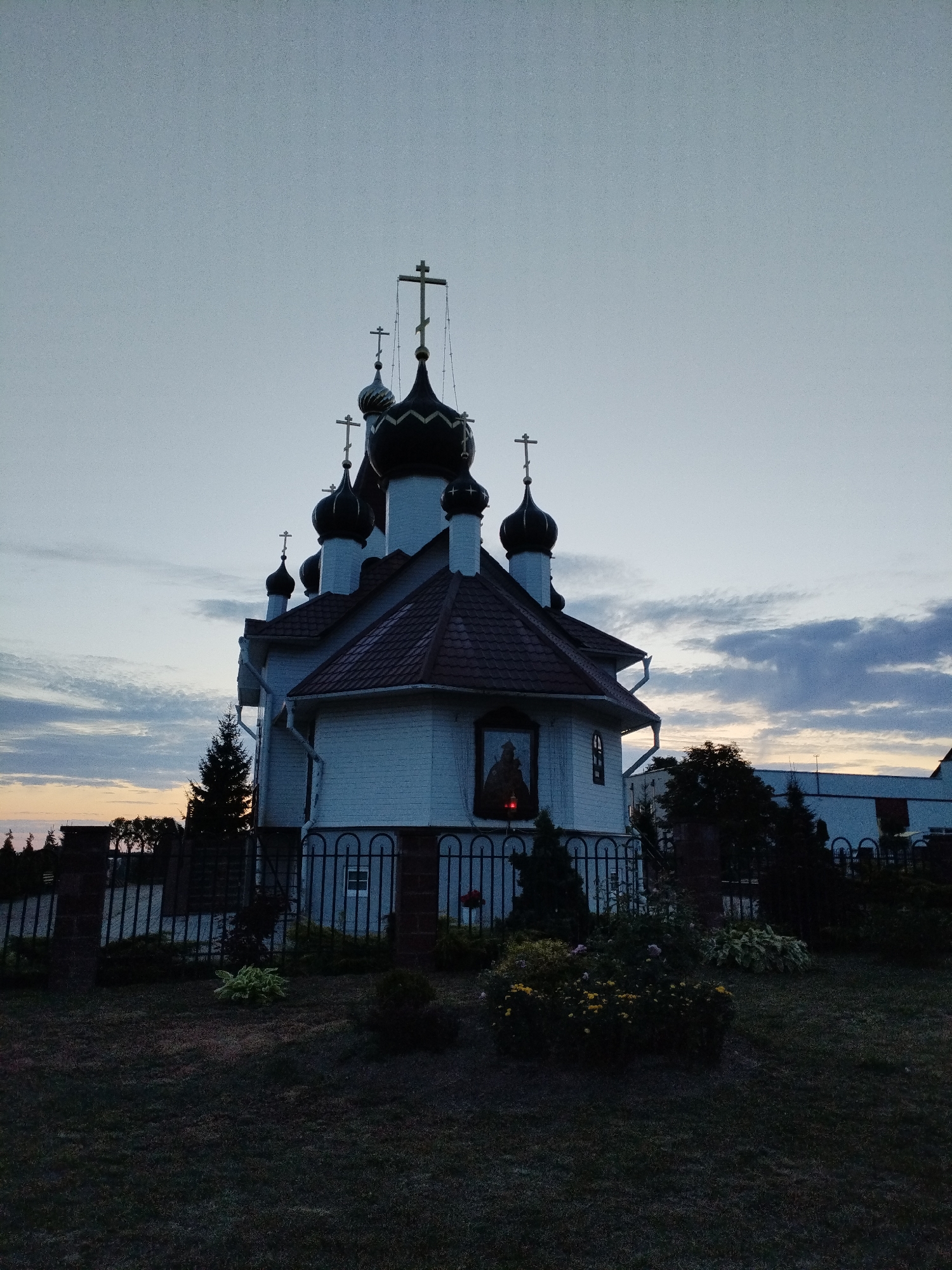 The image captures an Orthodox church in Bialynichy, Belarus, at dusk. The church is white with a red tiled roof and features multiple dark domes topped with crosses. A religious icon is visible in a window on the front of the building, with a red light beneath it. The church is surrounded by a black metal fence with brick pillars. In front of the fence, there are various shrubs and bushes, some of which are flowering yellow and pink. A tall evergreen tree stands to the left of the church. The sky is a pale gray with some wispy clouds, indicating the fading light of day. Other buildings and trees are visible in the background, silhouetted against the sky. The foreground is a dark grassy area.