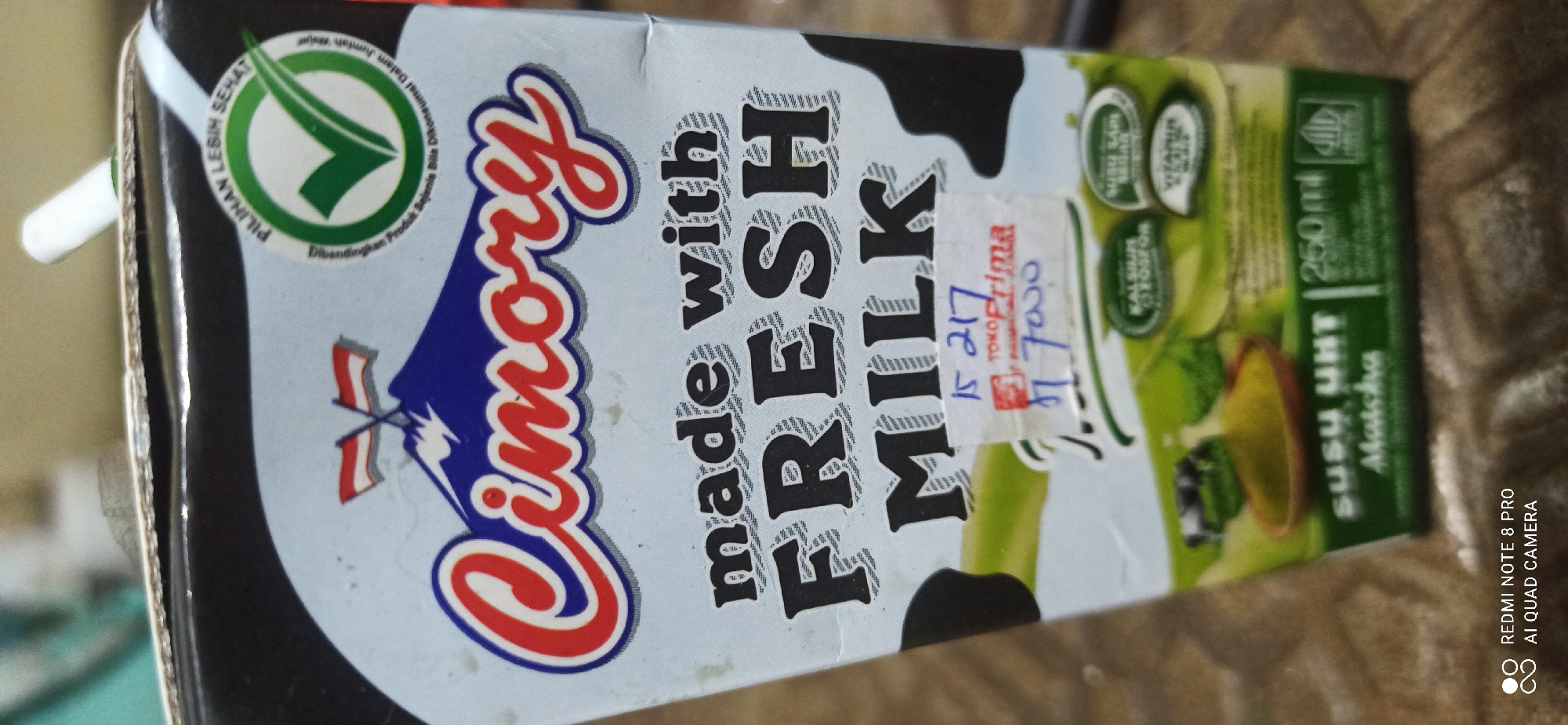 The image shows a close-up view of a carton of Cimory UHT Matcha flavored milk. The carton is light blue with black and red accents. The Cimory logo, featuring a mountain with two flags, is prominently displayed at the top. Below the logo, the text "made with FRESH MILK" is written in large black letters. A white sticker with handwritten text, possibly a price tag or inventory code, is attached to the front of the carton. In the lower section of the carton, there are green graphics depicting ingredients and nutritional information, including text such as "Kalsium Fosfor" and "Vitamin A". The background is blurred, but appears to be a textured surface. The image does not contain information about the location in Sedati Agung, Indonesia.