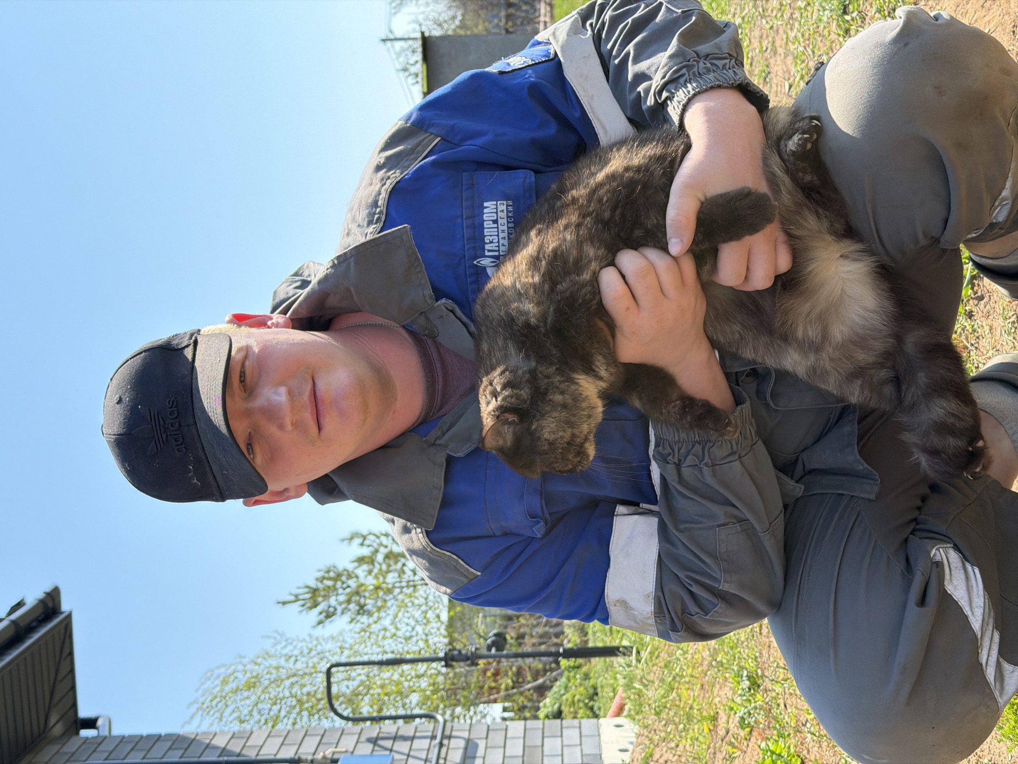 A man in a blue and gray Gazprom work uniform and a black Adidas baseball cap is holding a furry cat. He is sitting outside near a building. The location is Chaikovsky, Russia. The sky is blue.