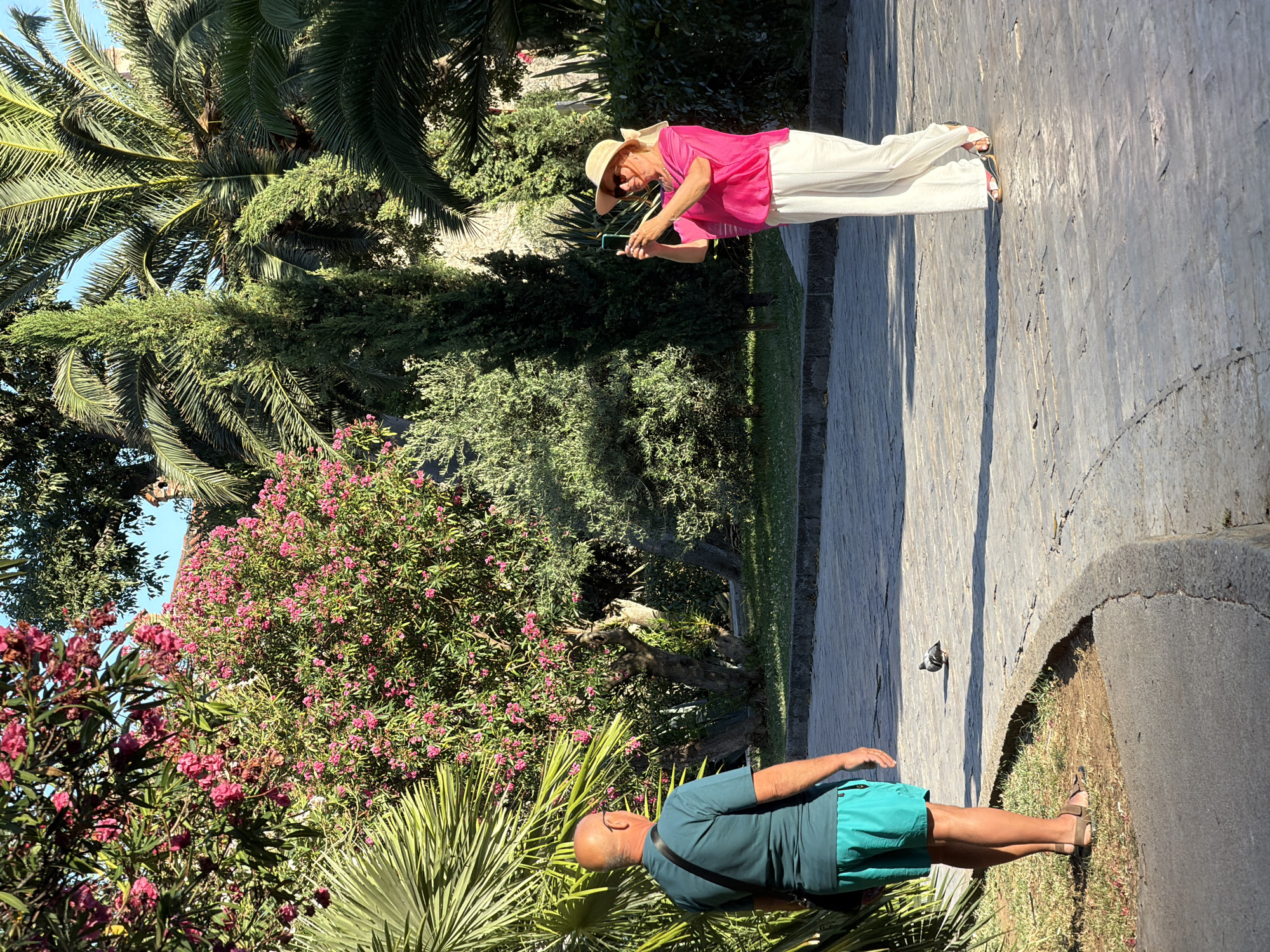Two people are standing on a stone-paved walkway in a park-like setting with lush vegetation. On the left, a bald man wearing a teal t-shirt and turquoise shorts stands facing away from the camera, with a dark bag slung over his shoulder. To the right, a woman wearing a wide-brimmed straw hat, sunglasses, a bright pink top, and white pants is holding up a smartphone, appearing to take a photo. A pigeon is visible on the walkway between the two individuals. The background features trees, including palm trees and flowering bushes with pink blossoms, suggesting a Mediterranean climate. The paved path curves gently, and grass borders the walkway. The lighting indicates it is daytime, with sunlight casting shadows. The scene is consistent with a public garden or park area within the city of Budva, Montenegro.