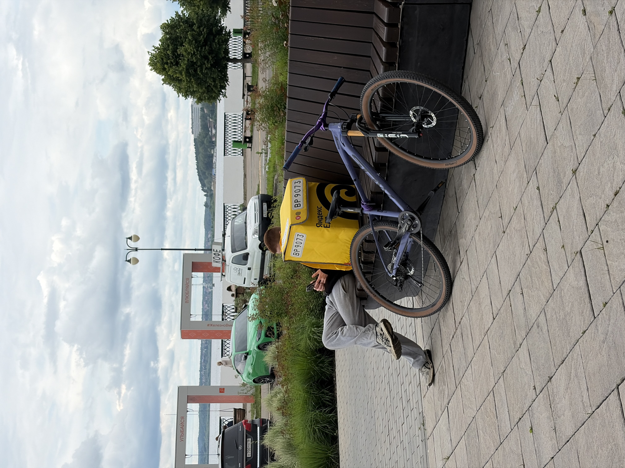 An outdoor, slightly rotated image captured in Izhevsk, Russia, depicts a person resting on a wooden bench beside a paved promenade overlooking a body of water. The scene takes place during the daytime under an overcast sky.
The primary subject is a person, likely a delivery courier, seated on the bench. They are dressed in grey sweatpants and a dark top, with a prominent bright yellow delivery backpack adorned with "Яндекс Еда" (Yandex Eda) and the number "BP 9073". The person is holding and looking at a smartphone. Leaning against the bench next to them is a purple mountain bike with a black suspension fork and tires marked "COMPASS".
In the background, a wide body of water (likely the Izhevsk Pond or Kama River) stretches towards a distant forested shoreline dotted with buildings. Along the promenade, there are various vehicles: a black SUV with license plate "O 700 TO 18", a green car (possibly a Kia Soul) with partially visible license plate "A033EY", and a white van. Metal railings line the waterfront, and a few trees and green bushes provide natural elements. A lamppost is also visible.
Several signs are present: two rectangular structures display the words "УЛЫБАЙСЯ" (Smile) and "ВЛЮБЛЯЙСЯ" (Fall in love), with a hashtag "#ЖелезноВместе" (IronTogether) partially visible on one. Another sign reads "КОФЕ" (Coffee). The ground is paved with light grey, rectangular tiles, with small weeds growing in the crevices. The overall impression is one of a brief rest taken by a courier in an urban park or waterfront area.