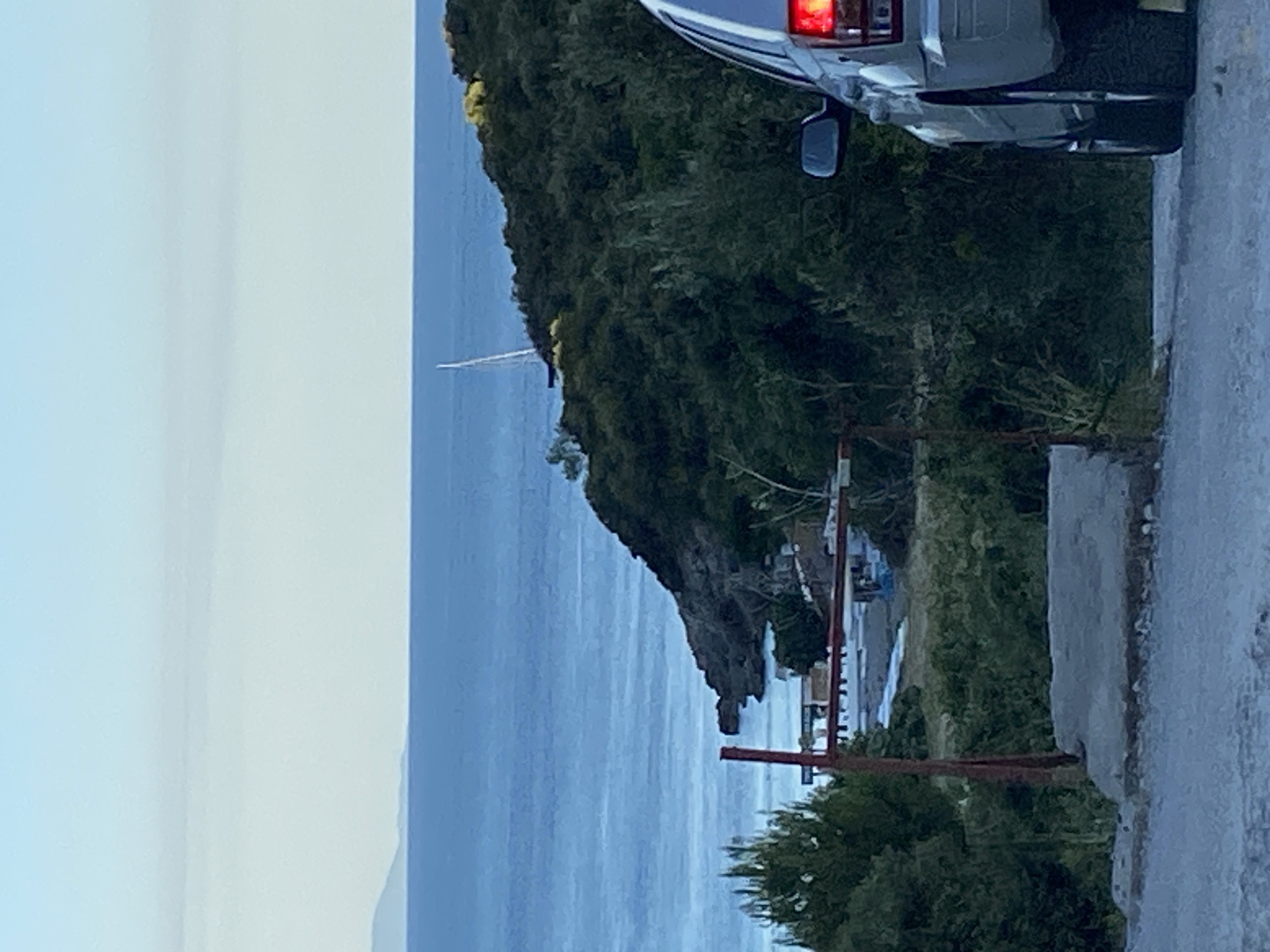The image depicts a coastal scene near Dubraljevina, Montenegro, under a clear, pale blue sky. The foreground features the rear of a silver SUV, parked on a gravel road. The SUV has its brake lights illuminated. A red metal structure, possibly a gate or fence, is visible to the left of the vehicle, partially obscured by greenery.
Beyond the immediate foreground, the landscape slopes down towards the sea. The sea itself is a deep blue, with a distant island or coastline visible on the horizon. A white sailboat with a prominent mast is positioned in the water, near a verdant, hilly area that juts out into the sea. This hilly area is covered in dense green foliage. At the base of the hill, some structures, possibly buildings or the remnants of a small settlement, are discernible. The overall impression is one of a quiet, natural setting, likely during the late afternoon or early evening given the soft light.