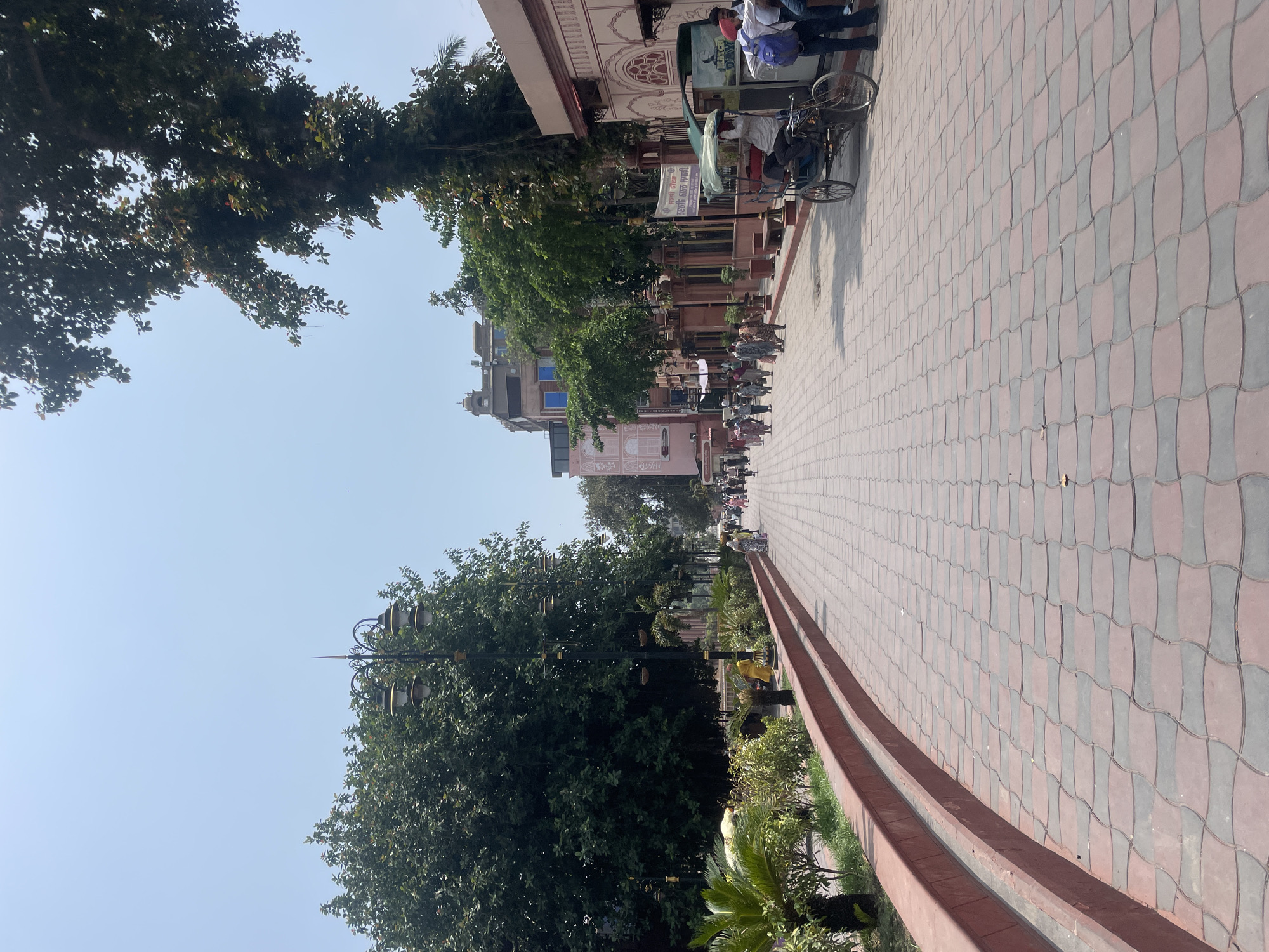 This is a photograph taken in Amritsar, India, likely depicting a street leading towards the Golden Temple, given the presence of a pedestrian walkway, religious architecture, and people dressed in traditional Indian attire. The image captures a sunny day with a clear blue sky.
In the foreground, a wide pedestrian walkway is paved with interlocking red and gray tiles, forming a curved pattern. The walkway is lined on the left by a raised red border with a planter box containing greenery and palm-like plants. Further left, large trees with dense foliage cast shadows. On the right side of the walkway, there are buildings with traditional Indian architectural elements, featuring arches and ornamental details, painted in light pink and white. A rickshaw, pulled by a man, is parked on the right side of the walkway, with a passenger seated under a canopy. Several people are walking along the pathway in both directions. Some are dressed in colorful traditional clothing, while others wear more casual attire. In the distance, more buildings and trees are visible. The overall scene suggests a vibrant public space within a city known for its religious and cultural significance.
