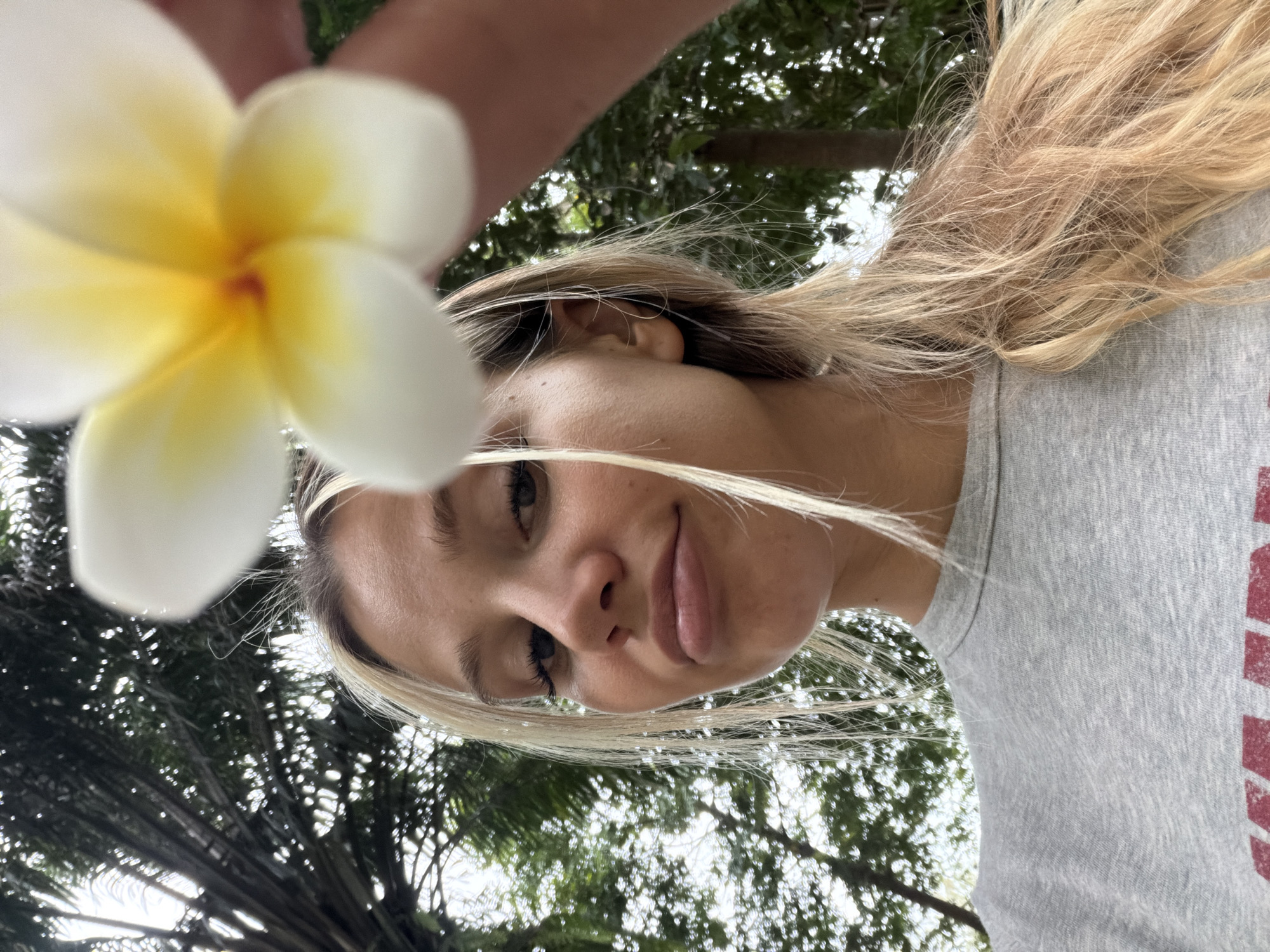 An extreme close-up selfie taken from a low angle captures a young woman with blonde hair and a grey t-shirt. A plumeria flower, out of focus, is held in the foreground, partially obscuring her face. She is looking directly at the camera with a slightly pursed expression. The background consists of out-of-focus greenery, suggesting an outdoor environment with trees. The scene appears to be in Choeng Thale, Thailand, indicated by the presence of palm trees common to the region.