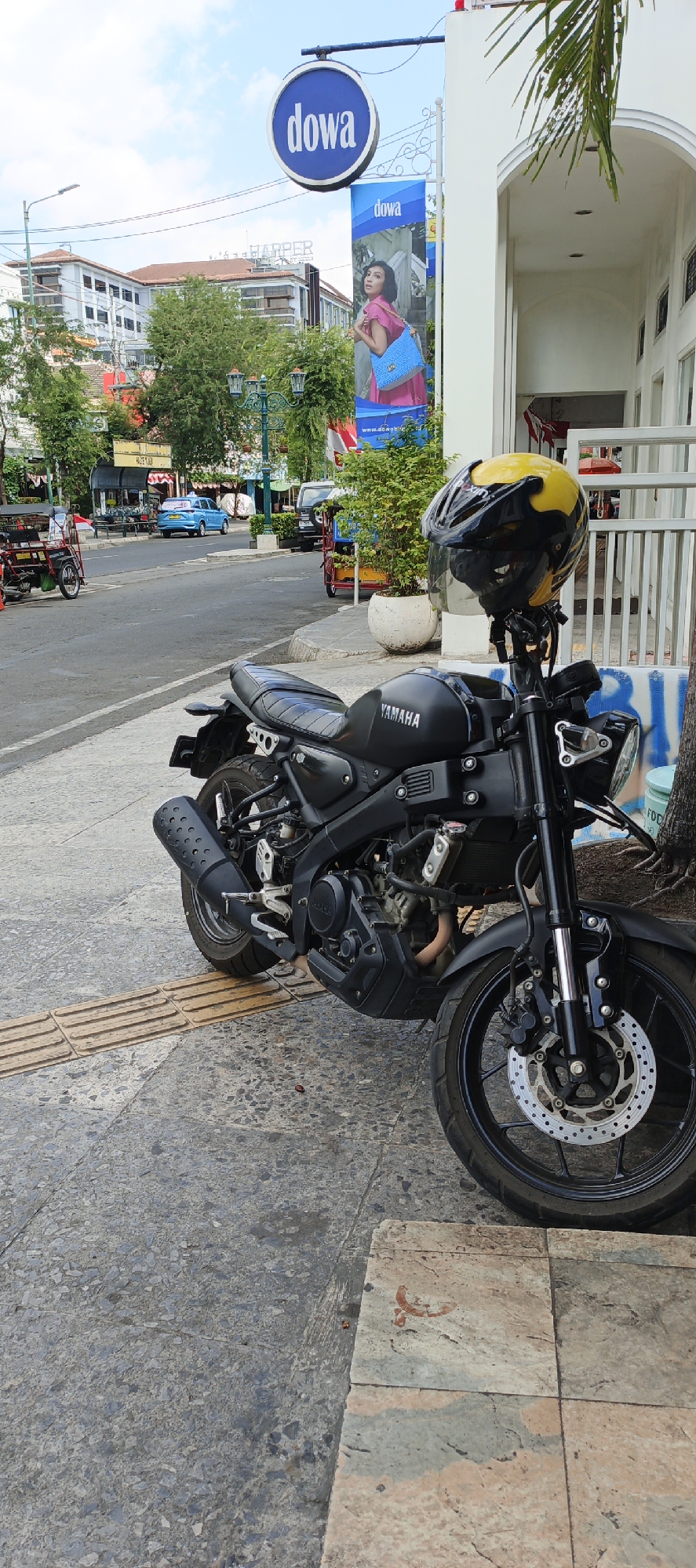 In Yogyakarta, Indonesia, a black Yamaha motorcycle with a yellow and black helmet is parked on a sidewalk. The motorcycle is positioned in the foreground, angled slightly towards the right. The sidewalk is paved with textured, rectangular tiles, with a raised tactile paving strip running along the edge. Behind the motorcycle, a street is visible with a blue car and a few motorbikes. In the background, there are buildings, trees, and a banner for "dowa" which is a shopping brand. The scene appears to be a commercial area with a mix of modern and traditional architecture. The sky is partly cloudy.