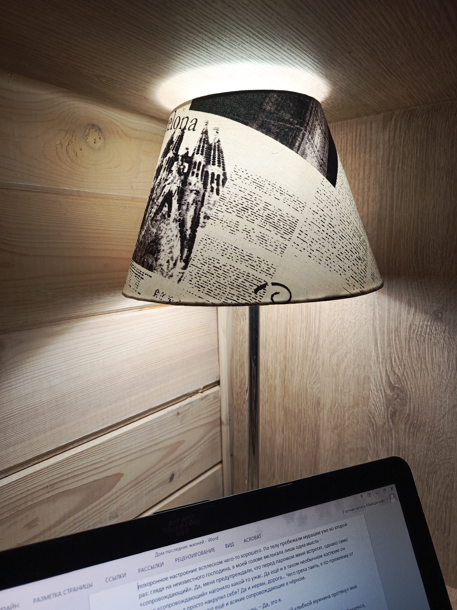 The image shows a room with wood paneling and a desk lamp with a shade that features a black-and-white pattern including the word "elona." The lamp sits between the wood panels of the room. The illumination from the lamp creates a bright spot on the ceiling above. In the foreground, a laptop screen displays a Word document with Russian text. The laptop appears to be in use, and the focus is drawn to the screen and the surrounding workspace. The city name mentioned is not visible.