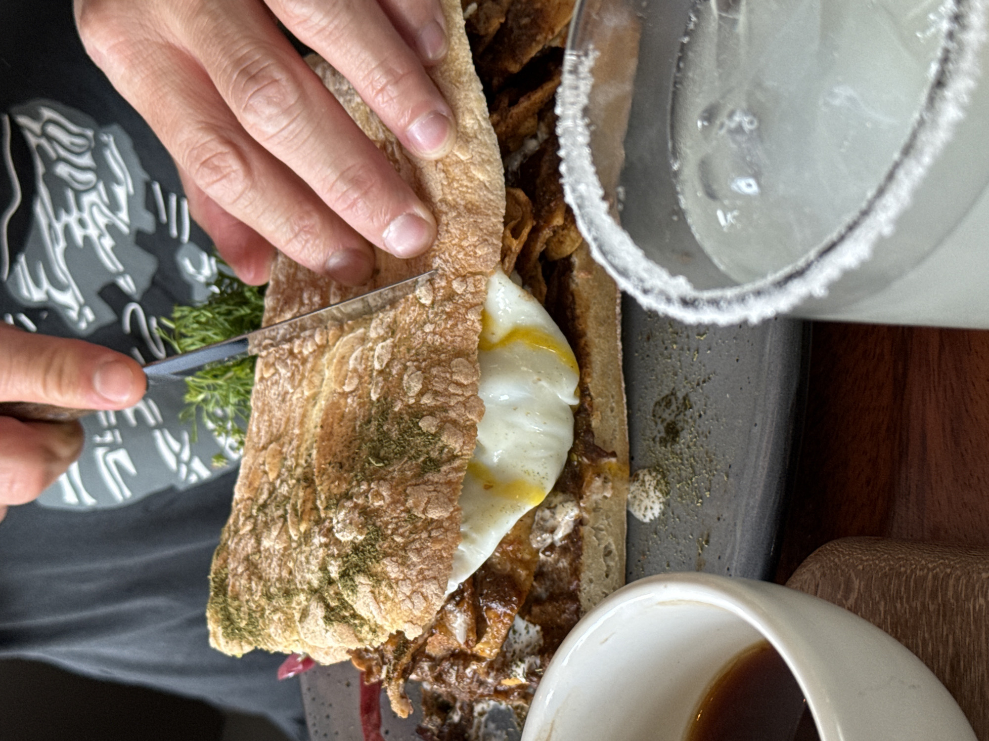 A person is cutting a sandwich with a knife. The sandwich appears to be on a dark gray plate, which also holds a small white bowl containing a dark liquid (possibly sauce or dressing). A glass with a sugared rim is positioned to the right of the plate. The person's left hand is holding the sandwich in place, while their right hand is cutting it with a knife. The background includes a wooden surface and a partially visible t-shirt with a white design. Based on research the location is likely in San Andrés Cholula, Mexico, though the exact establishment is not discernible from the image.