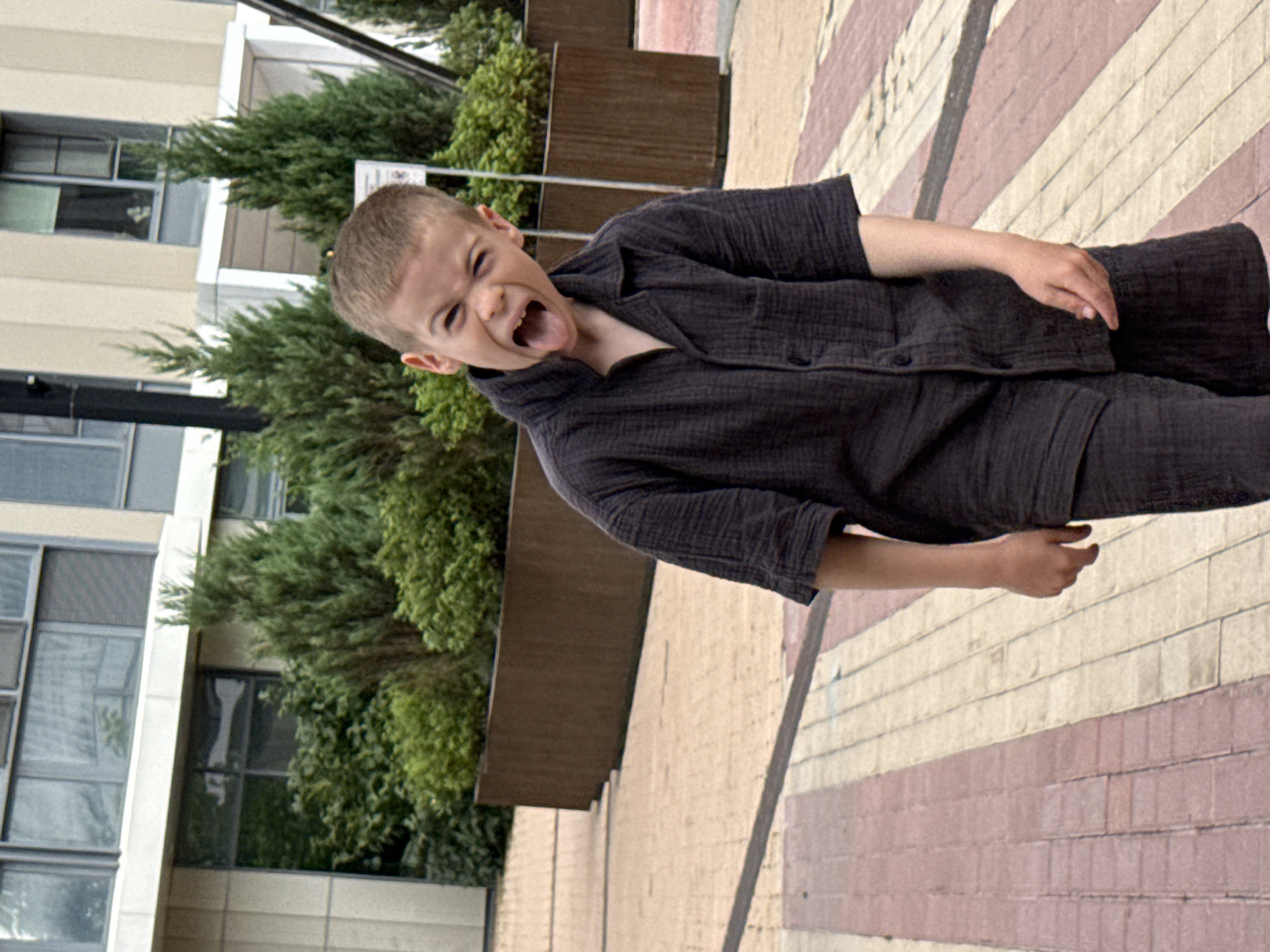 A young boy with short brown hair sticks his tongue out while facing the camera and stands on a patterned walkway made of pink and beige rectangular bricks in front of a modern building with large windows and lush green bushes. The boy is wearing a dark gray, short-sleeved collared shirt and matching shorts. The building appears to be residential or commercial, with a beige facade and multiple stories. The scene is set outdoors in an urban environment.