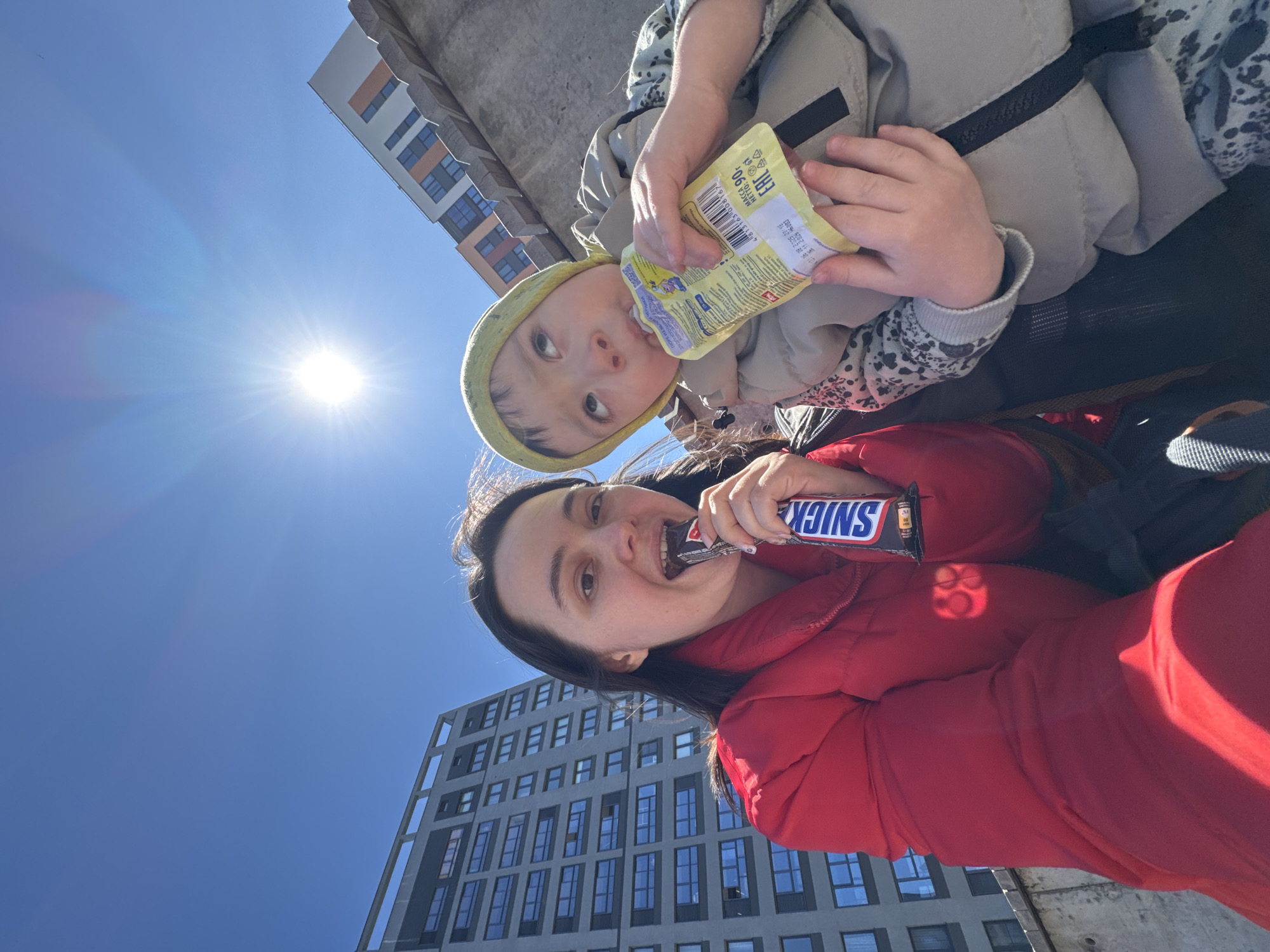 A selfie captures a woman and a young child outdoors in the city of Izhevsk, Russia. The woman is in the foreground, wearing a red puffer jacket, and eating a Snickers bar. Behind her, a modern high-rise building with many windows rises into the blue sky. The child, wearing a light green hat and light-colored jacket, is above and slightly behind the woman, drinking from a yellow juice pouch. The sun shines brightly in the upper left, creating a lens flare. Another building is also in the background.