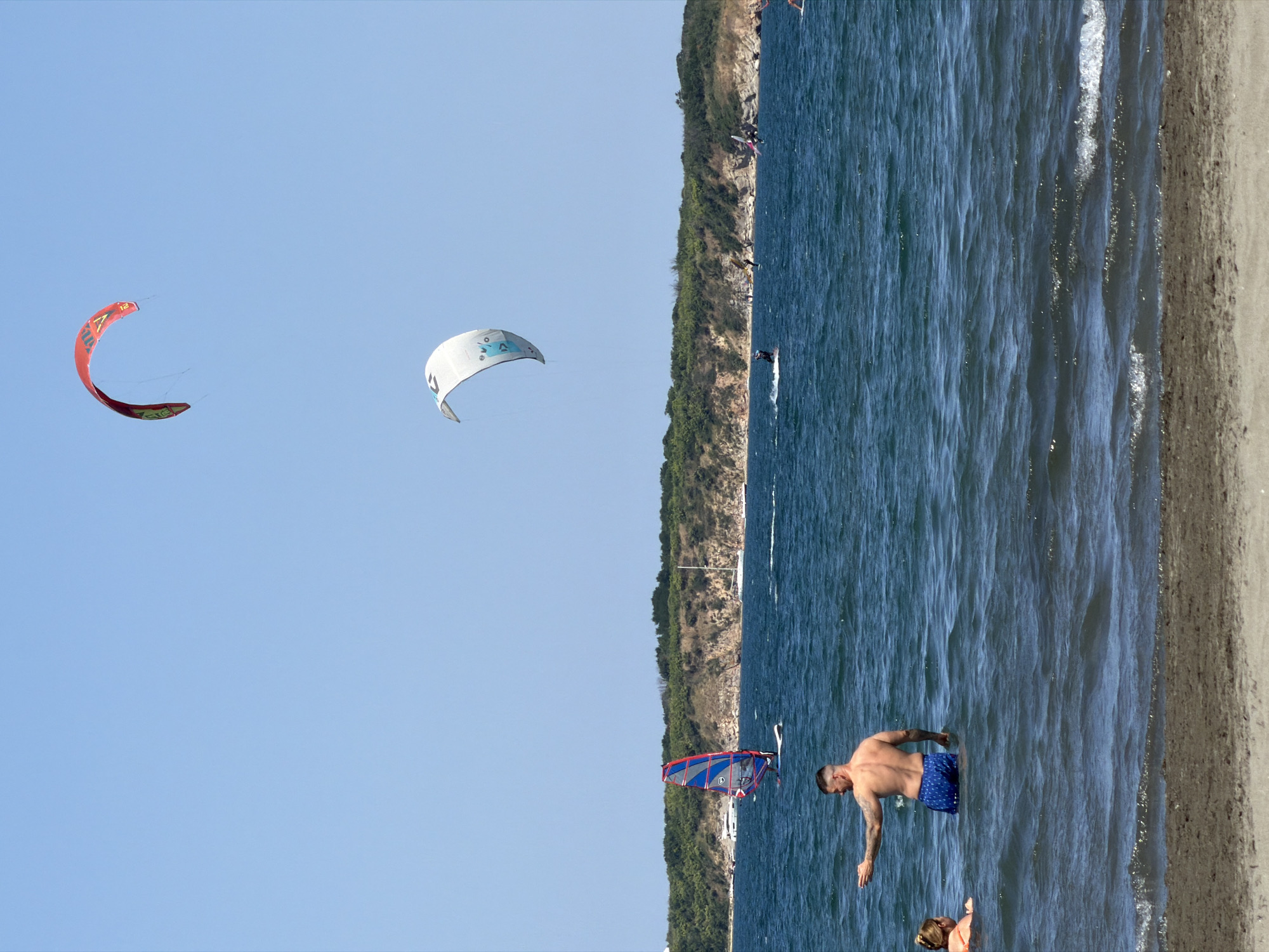 Atiya, Bulgaria, presents a vibrant coastal scene with various water sports activities under a clear blue sky. In the foreground, two individuals are partially submerged in the dark blue water, with a man in blue patterned swim trunks facing towards the left and gesturing with his right hand. A woman in an orange swimsuit is to his left, also in the water.
The middle ground showcases several windsurfers and kite surfers engaged in their respective activities. One prominent windsurfer with a red, blue, and white sail is visible. Further out, a white kite soars in the sky, and a red and yellow kite is also airborne. Numerous other individuals on boards are scattered across the water, indicating a popular day for water sports.
The background features a lush, green coastline with a rocky, eroded cliff face. A few small boats are moored near the shore, and a white sailboat is visible in the distance. The overall environment is that of a lively beach with a focus on recreational water activities.