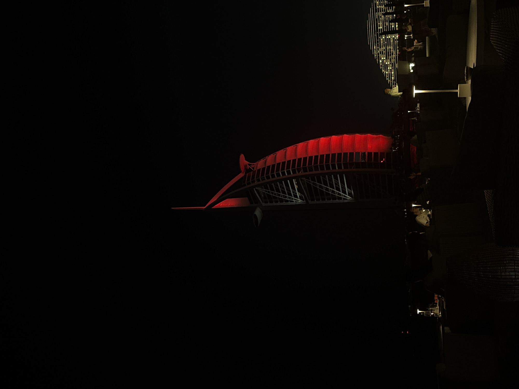 The image depicts a low-angle, night view of a section of the Dubai Water Canal Bridge in Dubai, United Arab Emirates. The bridge's central, elevated section is illuminated with red lights, casting a glow on its metal structure. Adjacent to the bridge is what appears to be a rooftop establishment, partially visible and illuminated with white lights, indicating the presence of people dining or socializing. The surrounding environment is dark.