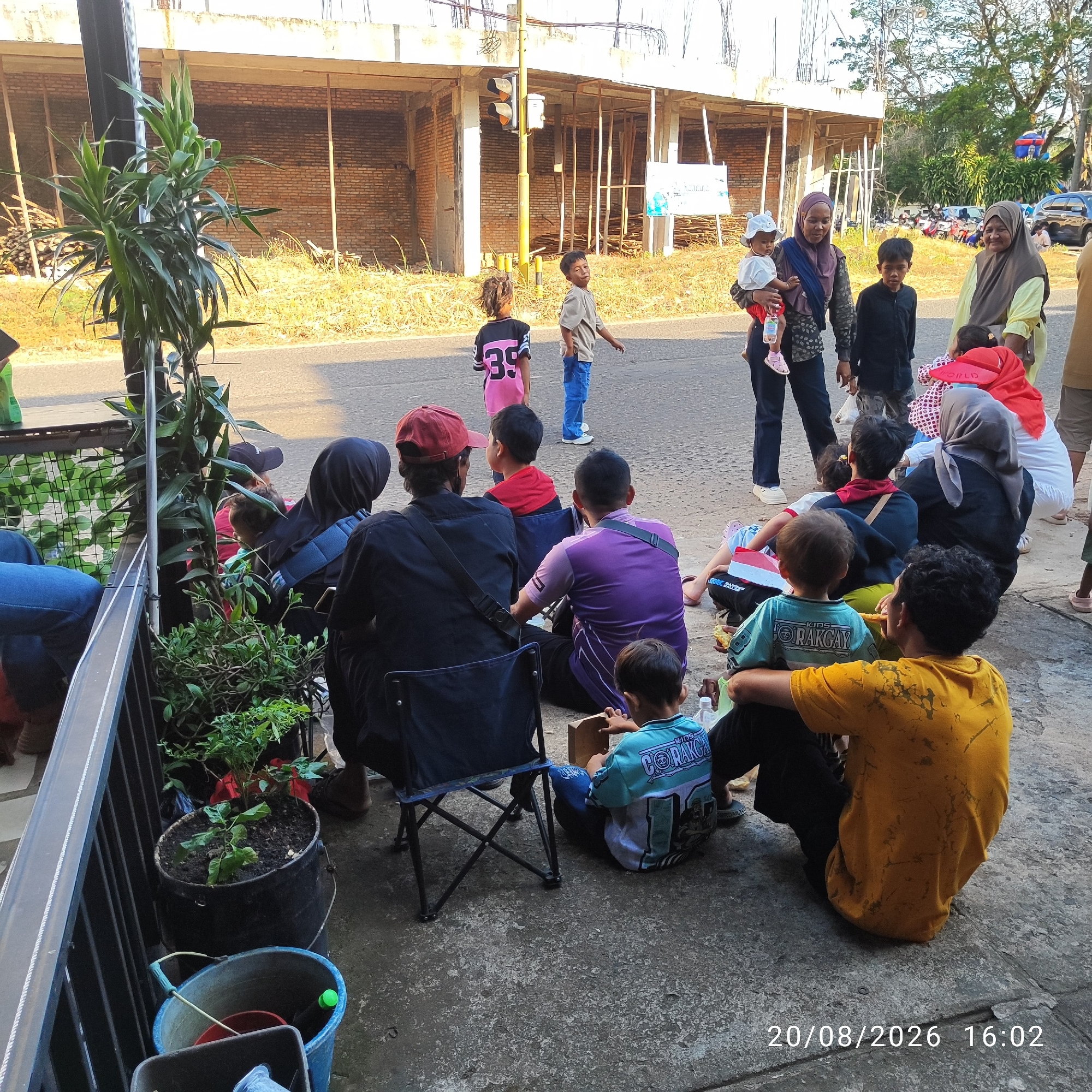 This is a street-level, eye-level, medium shot of a group of people, primarily women and children, gathered outdoors. The scene appears to be in Buding, Indonesia, suggested by the presence of a date stamp indicating "20/08/2026 16:02" and the general visual context.
In the foreground, several individuals are seated on the ground or in portable chairs, facing away from the camera. One person in a yellow t-shirt is seated with their back to the viewer. To their left, a child in a teal and white shirt is also seated. Next to them, a person in a purple shirt is sitting. Another person in a black shirt and a red cap is seated on the left, with their back to the camera.
Further back, a woman in a dark headscarf and blue jeans is holding a baby. To her right, a young boy in a black shirt stands. An older woman in a light yellow headscarf and a green shirt is also standing.
In the background, there are several children standing and interacting. One child in a pink and black shirt with the number "35" is facing away from the camera. Another child in a light brown shirt and blue jeans is standing and looking towards the left. A traffic light is visible on a pole in the background, along with scaffolding and the exterior of a brick building under construction.
The environment consists of a paved road and a sidewalk or dirt area where the people are gathered. There are also some potted plants and buckets visible in the foreground. The lighting suggests it is daytime, likely late afternoon, with sunlight casting shadows. The overall atmosphere appears to be a casual gathering.