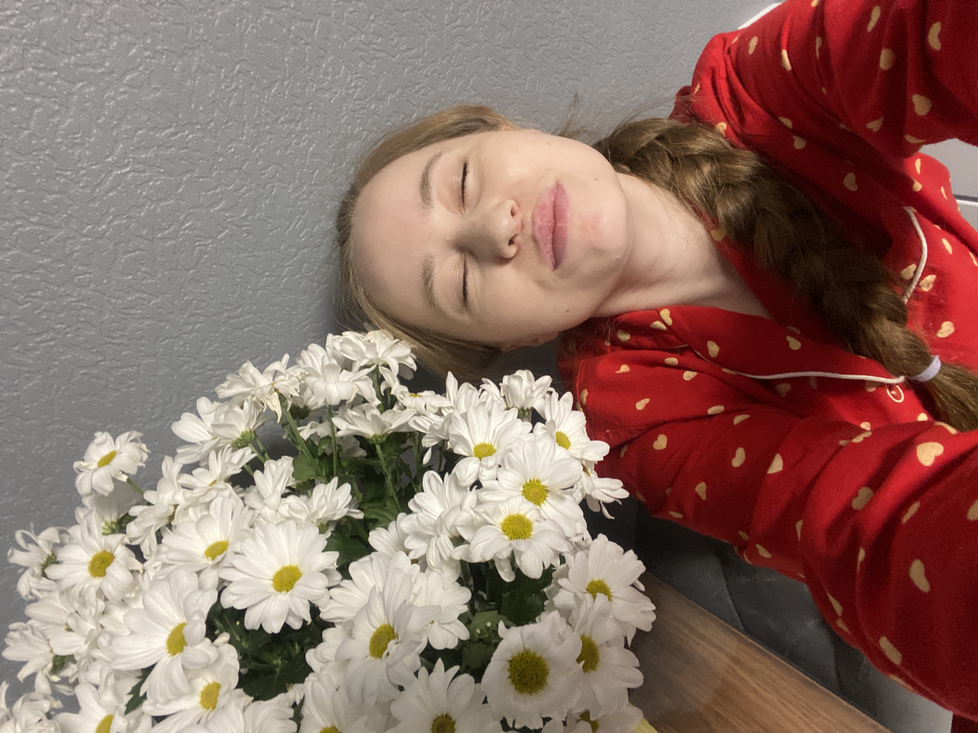 A serene, intimate selfie captures a young woman resting peacefully indoors, accompanied by a generous bouquet of white daisies. The woman, who has light brown hair styled in a braid, is wearing red pajamas patterned with numerous light-colored hearts. Her eyes are closed, and her lips are slightly parted, conveying an expression of deep relaxation or sleep. Her head is tilted and appears to be resting against a textured grey wall or headboard.
The vibrant white daisies, featuring bright yellow centers, are prominently displayed in the lower and central parts of the frame, creating a soft contrast with the woman's red sleepwear. A wooden surface is visible in the bottom right corner, beneath the flowers, while a darker grey or black surface, possibly part of furniture, can be seen behind her arm and alongside her. The overall mood is one of tranquility and comfort.
This moment of quiet repose is captured in Saint Petersburg, Russia. The time of day is not explicitly indicated, but the attire suggests either early morning or evening. No text is visible in the image.