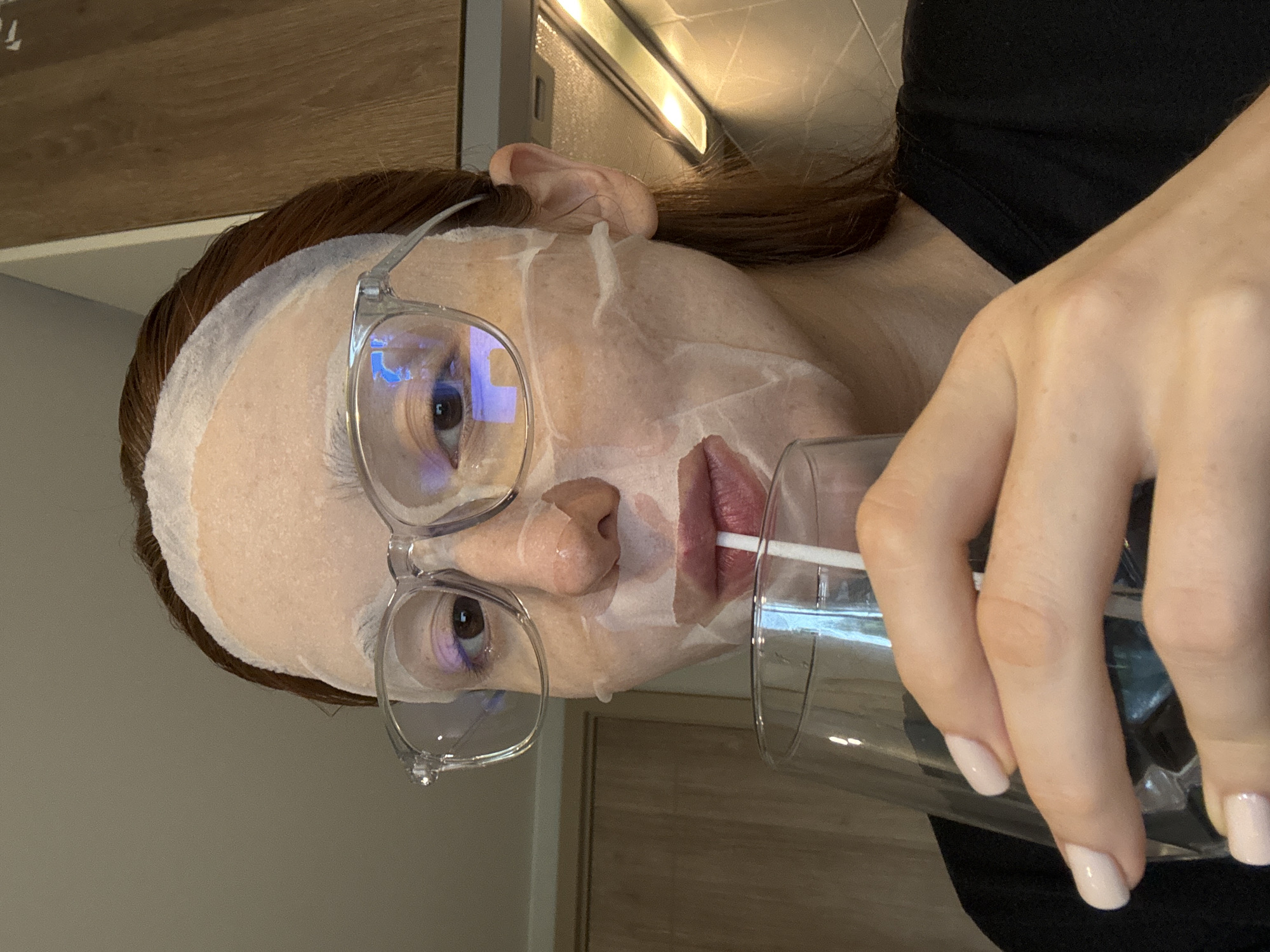 A woman with red hair and a sheet mask on her face is wearing clear-rimmed glasses and holding a glass with a straw. The location is potentially Choeng Thale, Thailand. The background includes what appears to be a cabinet.