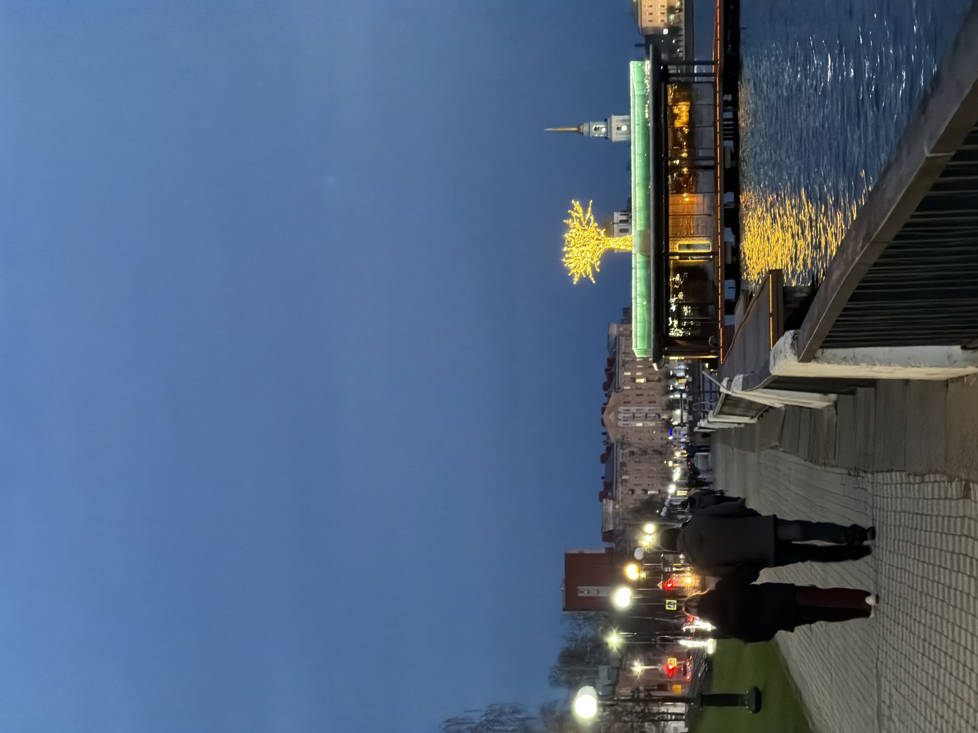 The image displays an urban waterfront scene in Votkinsk, Russia, captured during the evening or early night. The sky is a deep, clear blue, transitioning from dusk.
In the foreground on the left, a paved promenade or walkway extends into the distance. Several people are visible walking along this path, with two individuals in dark attire prominently silhouetted against the lights in the mid-ground, appearing to stroll away from the viewer. Streetlights line the promenade, casting light on the patterned surface.
To the right, a body of water, likely a river or canal, stretches across the frame, reflecting the vibrant city lights in shimmering streaks of yellow and white. A dark railing separates the pedestrian path from the water.
Across the water, various buildings are illuminated, contributing to the urban glow. A striking yellow, tree-shaped light installation stands out brightly on the opposite bank. Behind it, a building with a distinctive, slender spire rises against the twilight sky. Another building on the near side features unique bright green illuminated elements along its facade, adding to the city's evening illumination.
In the further distance on the left, red and yellow traffic light signals indicate the presence of a road and vehicle traffic beyond the pedestrian area. The overall atmosphere appears calm and pleasant, with the abundant artificial lighting creating an inviting and possibly festive ambiance. There is no discernible text visible in the image.