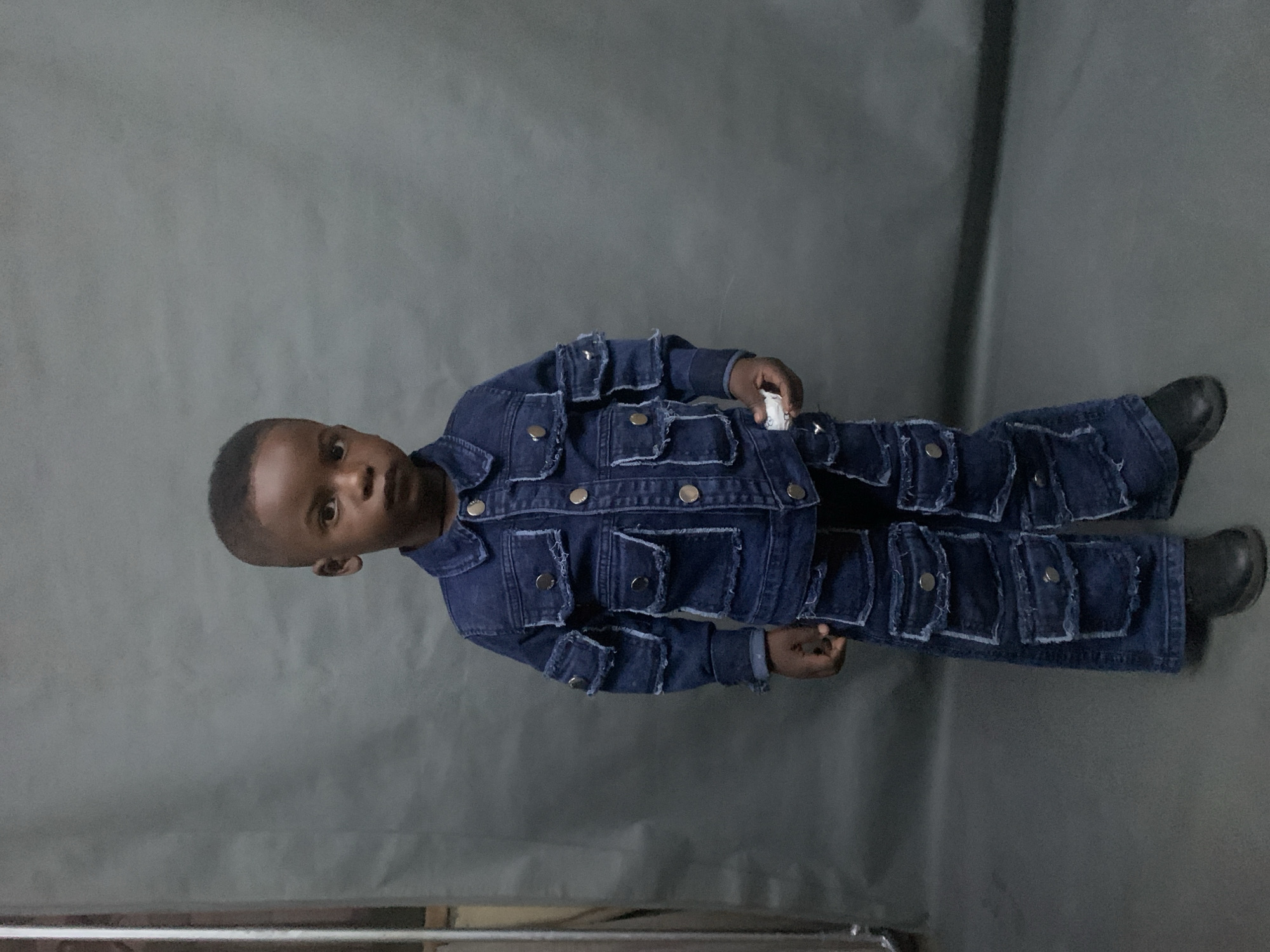 A young boy is lying on a gray surface. He is wearing a dark blue denim outfit with numerous pockets and silver buttons. He is wearing black shoes. He is holding a small white object in his right hand. The location is unknown; there is no indication of it being Jos, Nigeria.
