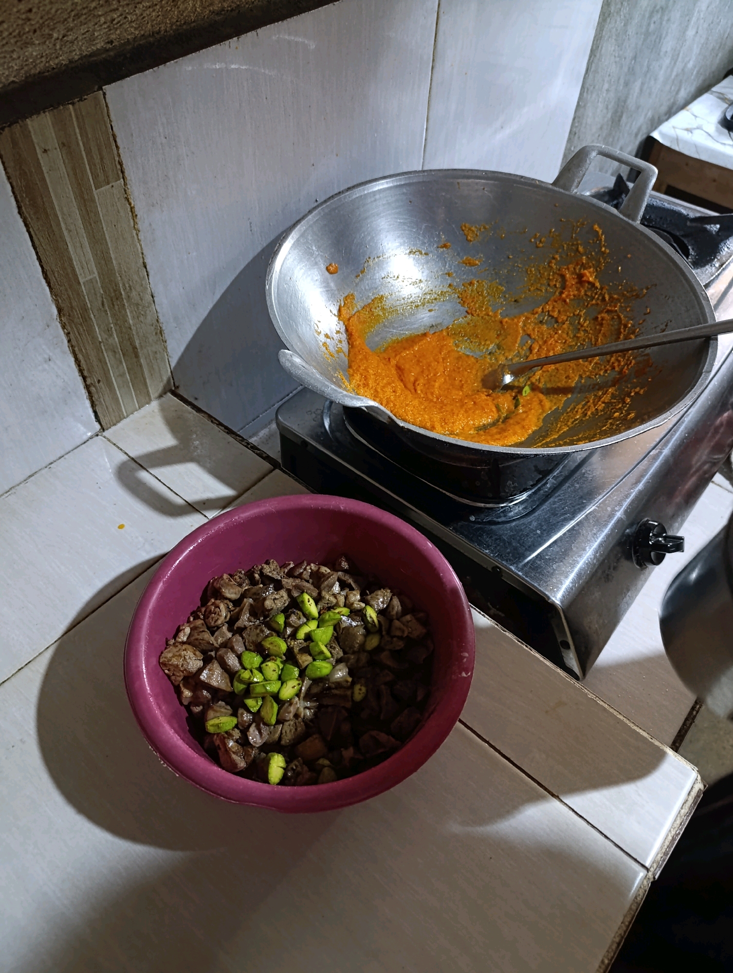A dark, overhead shot captures a kitchen scene in a modest dwelling, possibly located in the city of Bandung, Indonesia. A well-used aluminum wok, containing a vibrant orange spice paste with hints of green, sits atop a gas stove. To the left, a deep, magenta bowl is filled with diced, dark brown meat, possibly liver, interspersed with bright green pieces of petai (stink beans). The tiled countertop and backsplash suggest a practical, lived-in kitchen environment. The lighting casts long shadows, hinting at the time of day, possibly evening. There are no people visible in the frame.