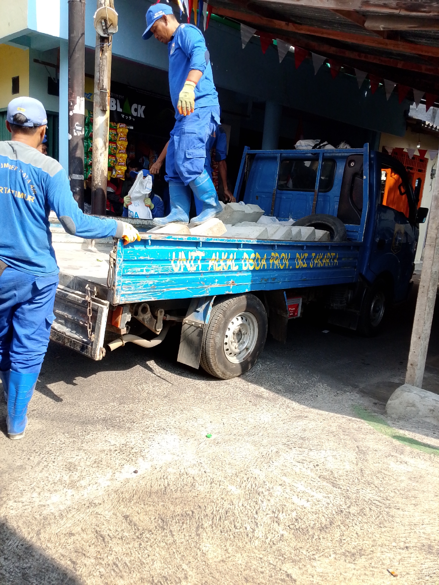 Two individuals wearing blue uniforms and caps are engaged in loading concrete blocks onto the back of a blue utility truck. One individual, facing away from the camera, is standing on the edge of the truck bed, holding a white bag. The second individual, also in uniform, is standing on the ground next to the truck, reaching towards the white bag. The truck is parked on a textured concrete surface. In the background, a building with a blue facade is visible, along with what appears to be a storefront displaying various products on shelves. Red and white triangular flags are strung across the top of the scene. The scene suggests a loading or unloading operation taking place in an outdoor urban environment, likely in East Jakarta, Indonesia, given the markings on the truck which read "UNIT ALKAL BSDA PROV. DKI JAKARTA".