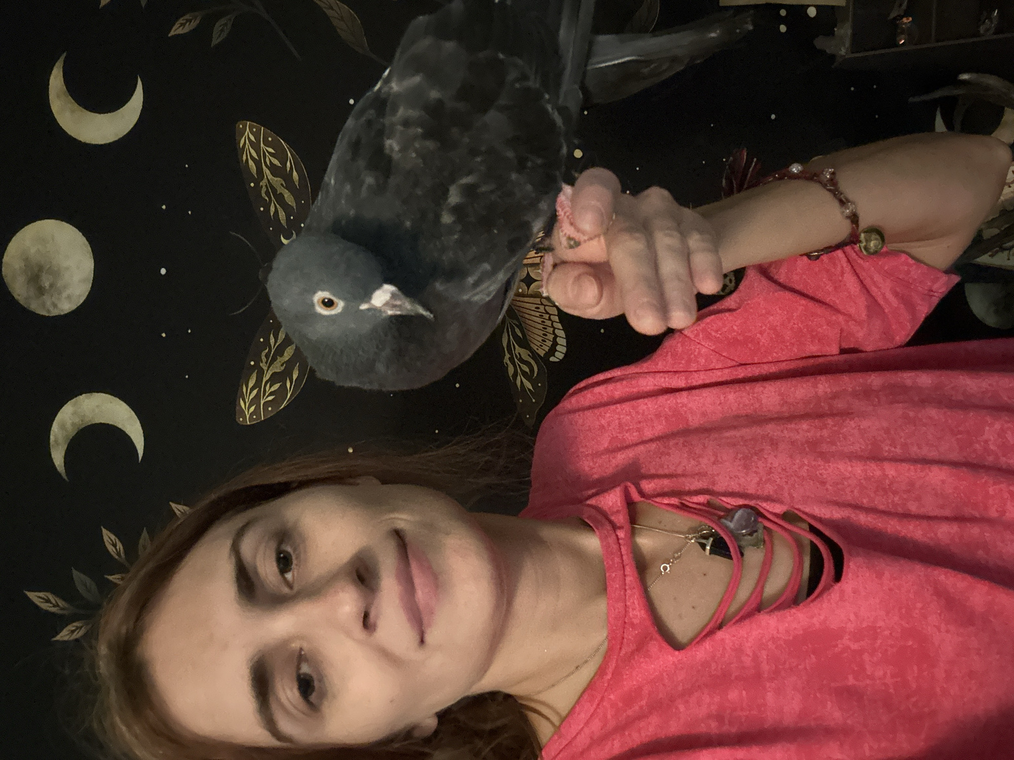 A woman with reddish-brown hair and brown eyes is holding a pigeon on her hand. She is wearing a bright pink shirt with cutouts on the chest and a necklace with several pendants. The background is a black wall decorated with celestial motifs, including moons and moths. The scene is indoors and appears to be a personal space. There is no information about the location in Togliatti, Russia.
