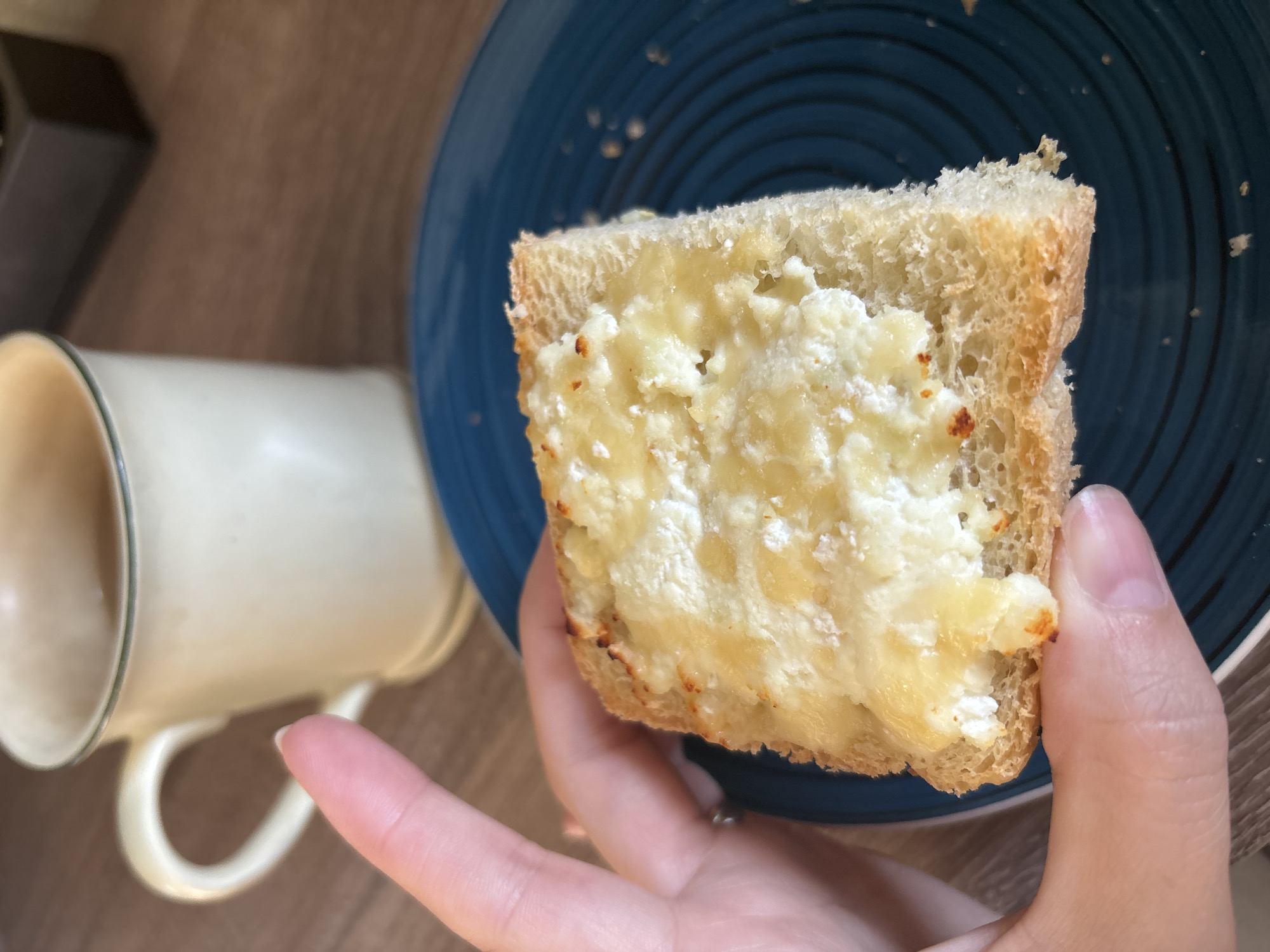 A close-up, eye-level shot shows a hand holding a slice of toast topped with melted cheese, resting on a dark blue plate. To the left of the plate, a light-colored mug with a green rim contains a brown beverage, likely coffee or tea. The background appears to be a wooden surface, possibly a table. The scene is indoors, suggesting a casual meal or snack. There is no visible indication of the specific location within Naberezhnye Chelny, Russia.