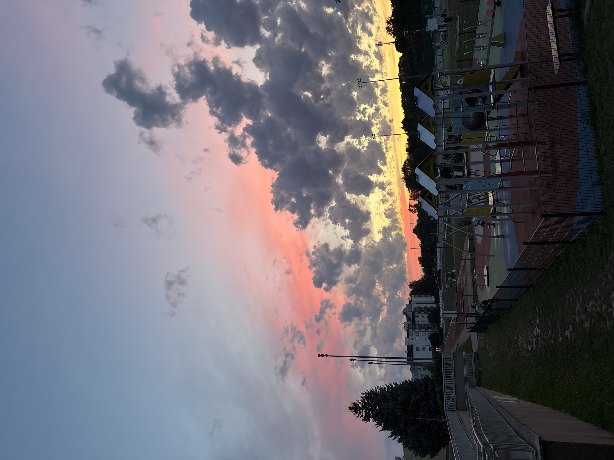 The image depicts a playground and a sports field at dusk, with a vibrant sky painted in shades of pink, orange, and blue, illuminated by the setting sun. The playground features colorful slides, swings, and climbing structures, with a few children visible playing. Adjacent to the playground is a sports field, possibly a football pitch, with floodlights and some spectators. In the background, residential buildings and trees are silhouetted against the colorful sky. The scene appears to be a public space in Lublin, Poland, given the context provided.