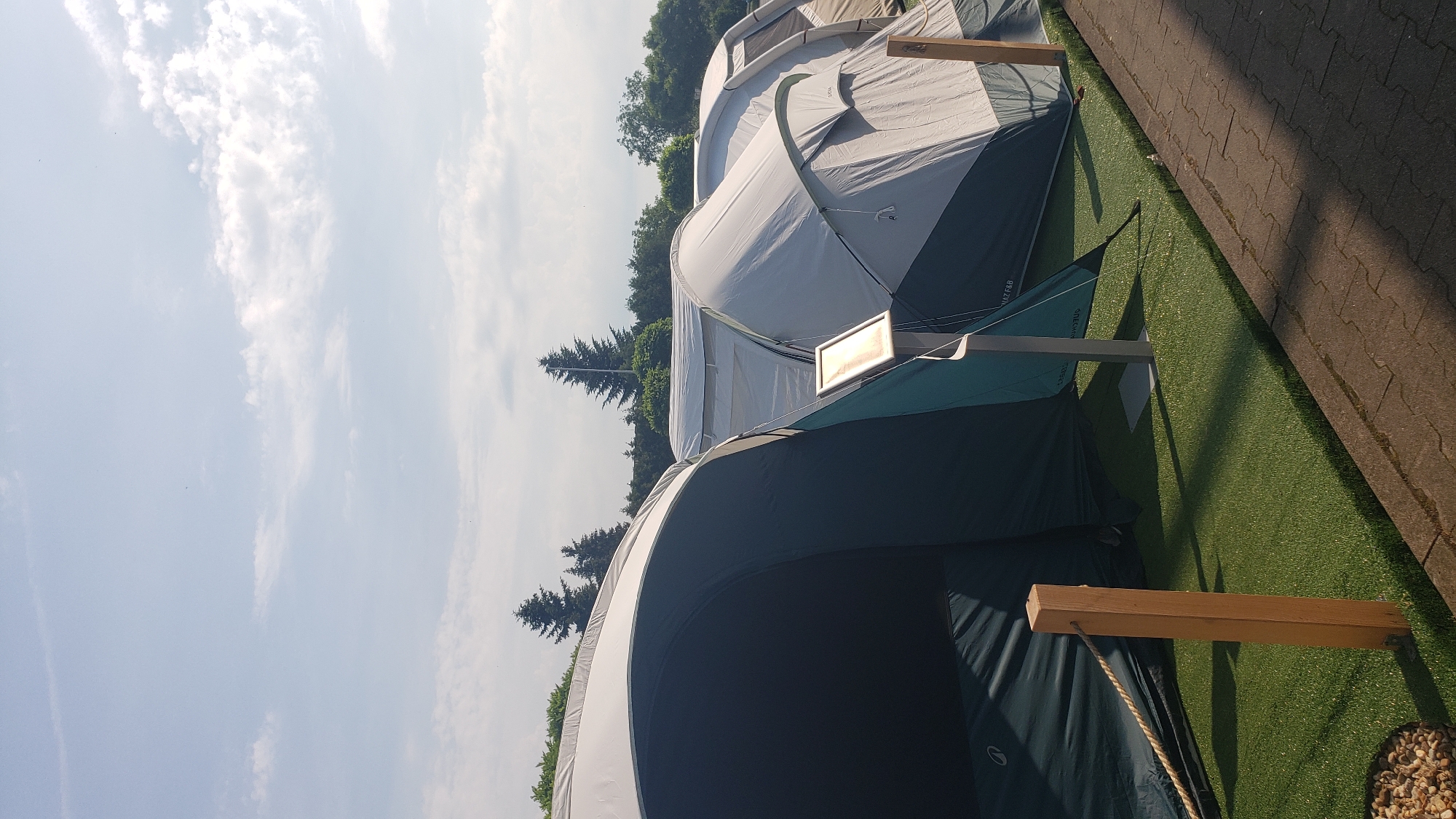 The image shows a campsite with several tents set up on artificial grass. The tents are of various sizes and colors, with a large dark green and white tent in the foreground, followed by several grey and white tents. There are wooden posts and rope barriers along the edge of the campsite. In the background, there are trees and a cloudy blue sky. The scene appears to be a typical outdoor campsite, possibly located in or near the city of Prešov, Slovakia. There are no people visible in the image.