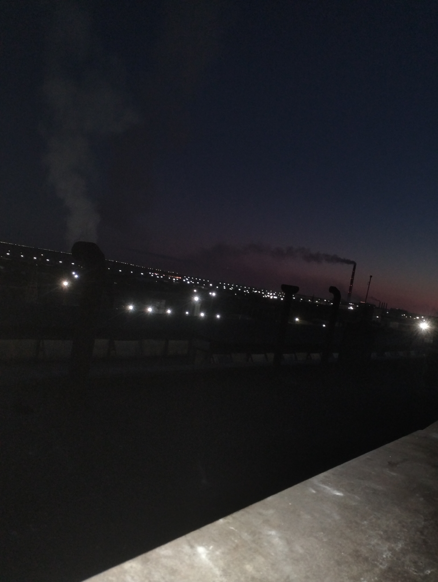 The image captures a nighttime industrial landscape in Jezqazğan, Kazakhstan. In the foreground, a dark, silhouetted structure, possibly a railing or a wall, runs diagonally from the bottom right to the left edge. Behind it, a darker, indistinct area suggests a foreground building or platform.
The middle ground features a series of illuminated industrial structures and elements. Several tall, dark vertical shapes are visible, some with unusual curved tops. A notable feature is a tall, reddish-orange smokestack emitting a plume of dark smoke into the dusky sky, suggesting ongoing industrial activity. Scattered across the middle ground are numerous bright, white lights, likely streetlights or facility lights, creating a dotted pattern against the dim surroundings. A lighter plume of smoke or steam rises from a more distant, dark vertical structure on the left.
The background is dominated by the dark night sky, which transitions to a faint reddish hue near the horizon, possibly indicating the last vestiges of sunset or light pollution. The horizon itself is marked by a dense arrangement of lights, suggesting a town or city in the distance. The overall atmosphere is one of industrial activity at night, with artificial lights illuminating the scene against the dark sky.