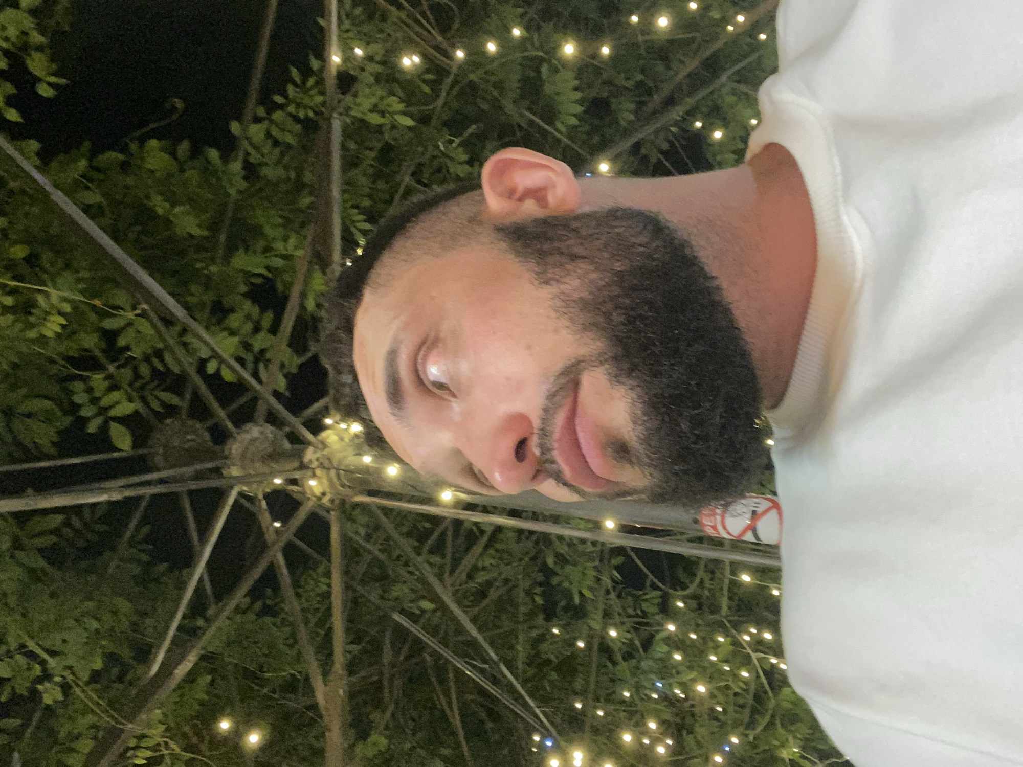 The image shows a man with a beard and short hair, wearing a white sweatshirt. He is outdoors at night, with a canopy of trees and string lights above him. The scene appears to be in an outdoor seating area, possibly a cafe or restaurant. The man is looking up and to the right, with his face illuminated by the ambient light. In the background, there are more string lights and the dark silhouettes of trees and branches. There is also a "No Smoking" sign visible on a pole behind the man. There is no indication that this location is in Makhachkala, Russia.