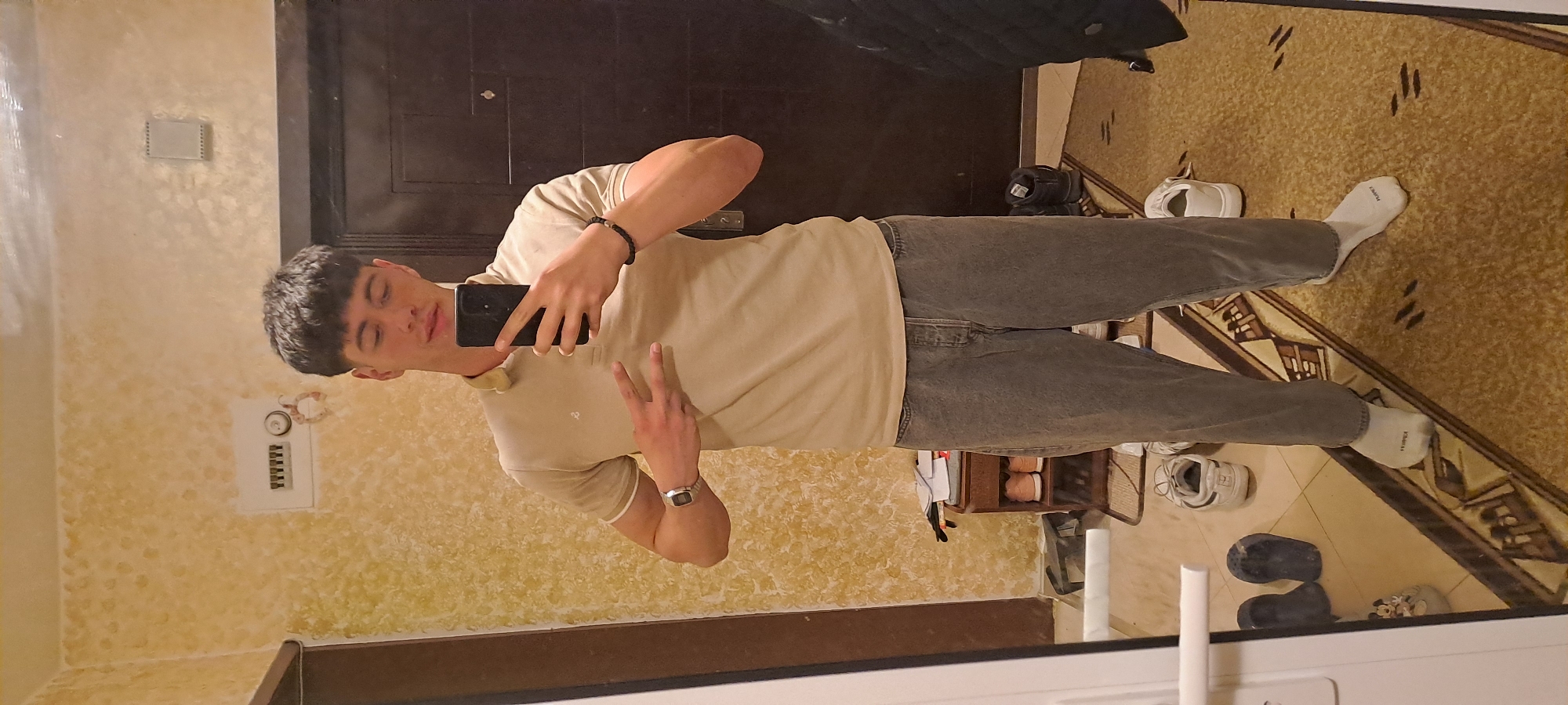 A person stands in front of a mirror, likely indoors. The individual is wearing a beige polo shirt, grey jeans, and white socks. They are holding a black smartphone and making a peace sign with their left hand. A watch is visible on their left wrist, and a beaded bracelet on their right wrist. The background includes a dark-colored door, a patterned rug, and a wallpapered wall. Footwear is scattered around. The location is unknown, but it could be Burgas, Bulgaria.